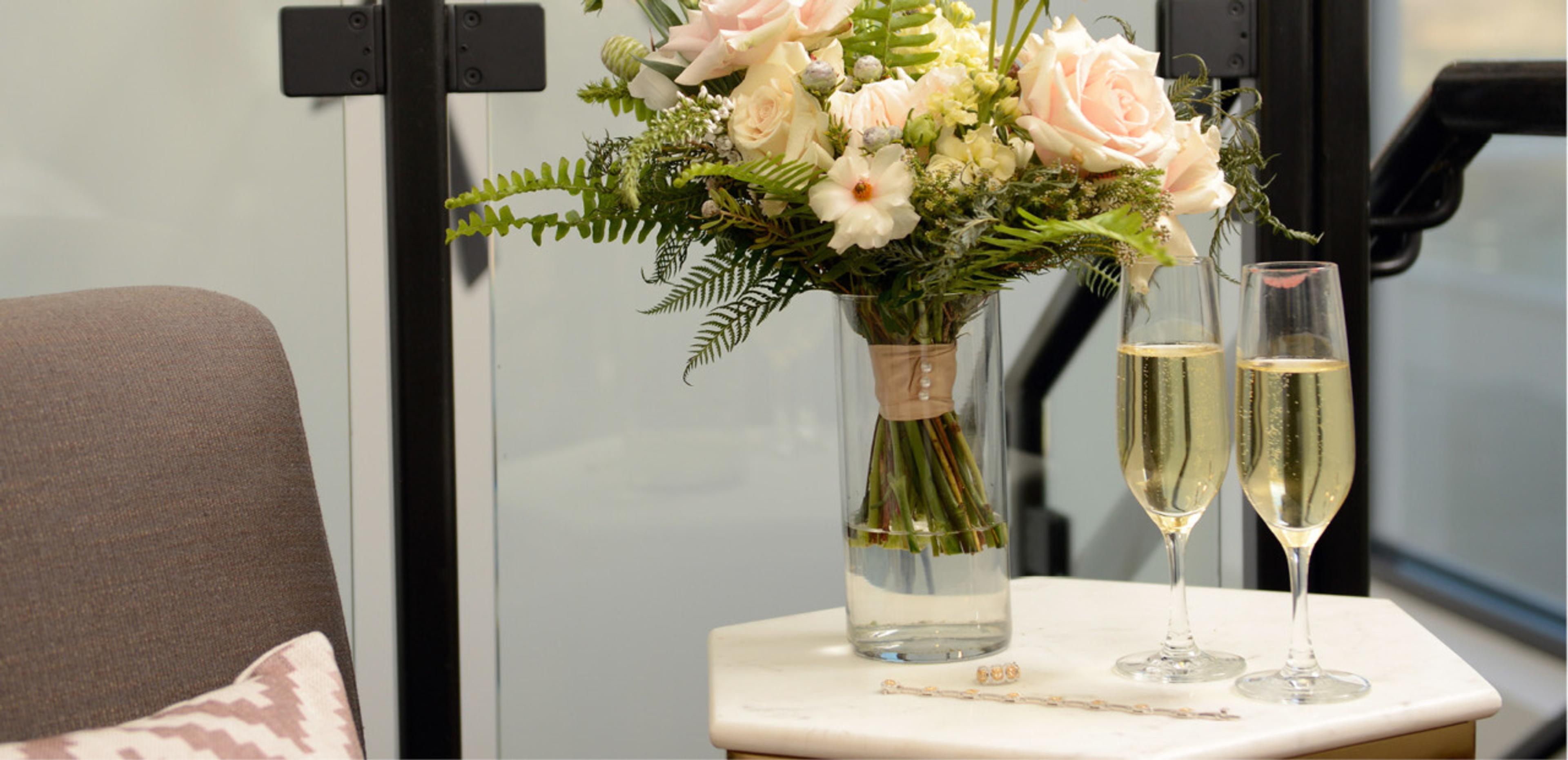 Pink and white flowers in a clear glass vase and two glasses of champagne sit on a white marble side table beside a glass staircase and a grey armchair.