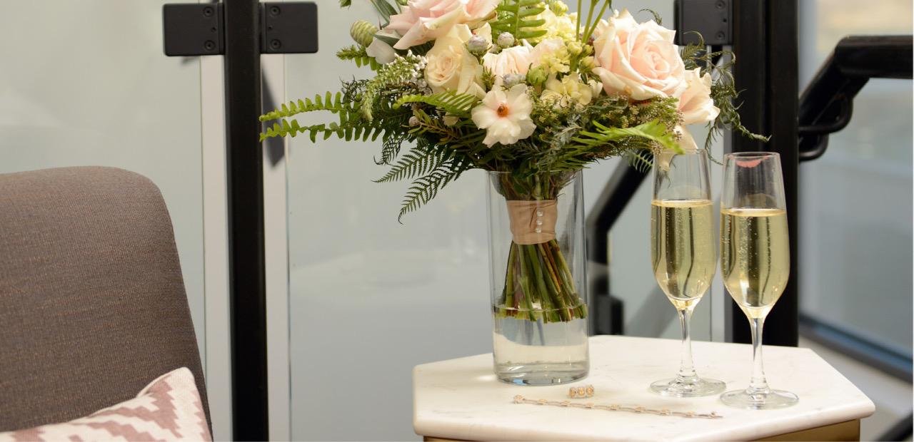 Pink and white flowers in a clear glass vase and two glasses of champagne sit on a white marble side table beside a glass staircase and a grey armchair.