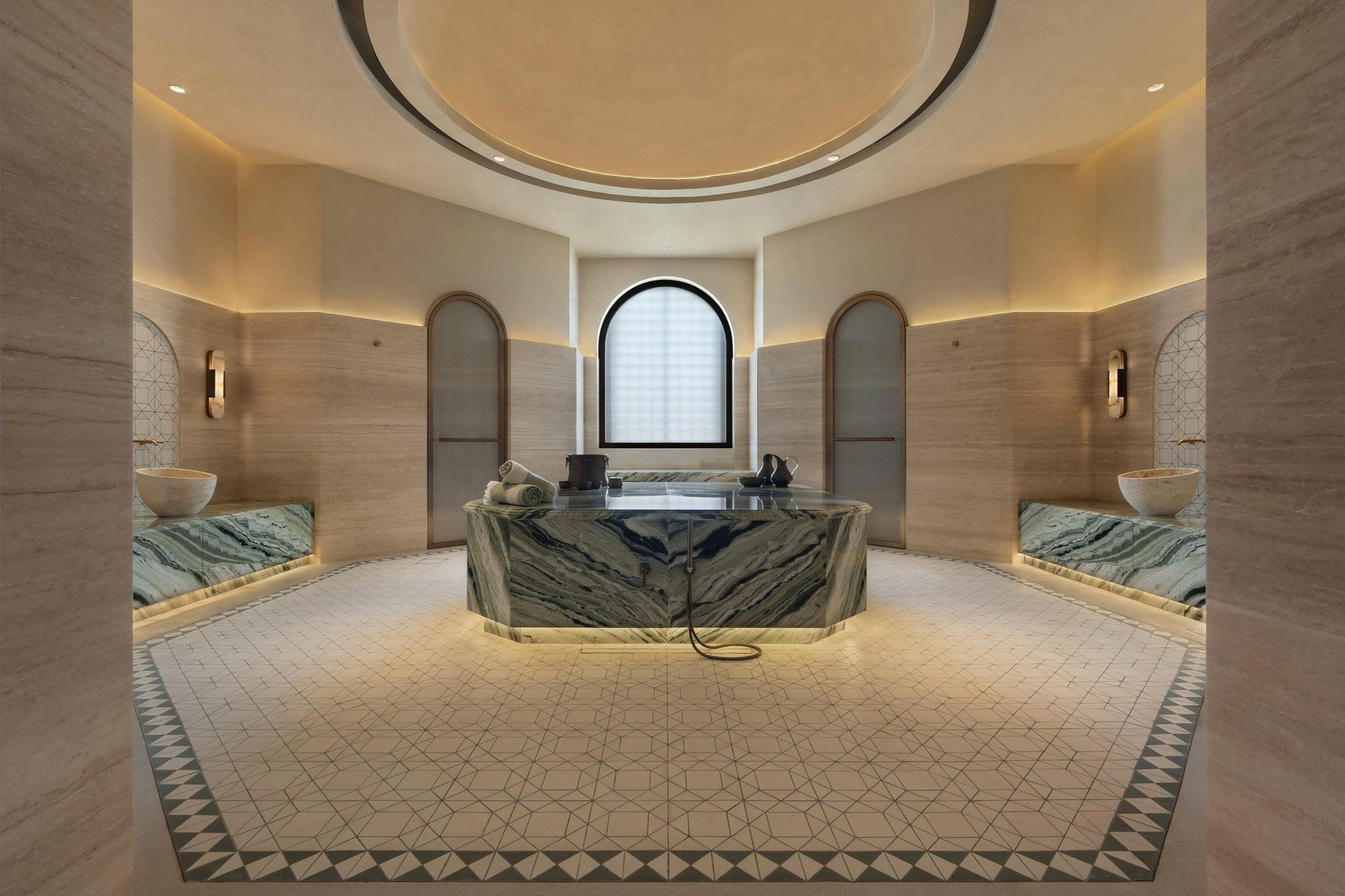 An empty marble hammam room with a central heated stone slab, arched doorways, and a domed skylight.