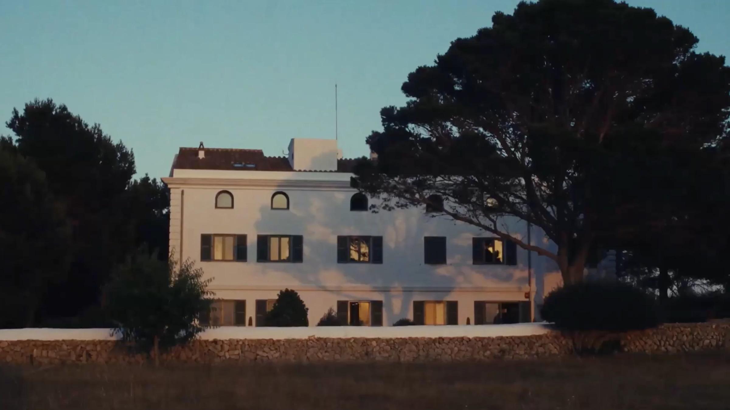 A white three-storey building with shuttered windows and a low stone boundary wall, lit by the low sun among tall pine trees at dusk.