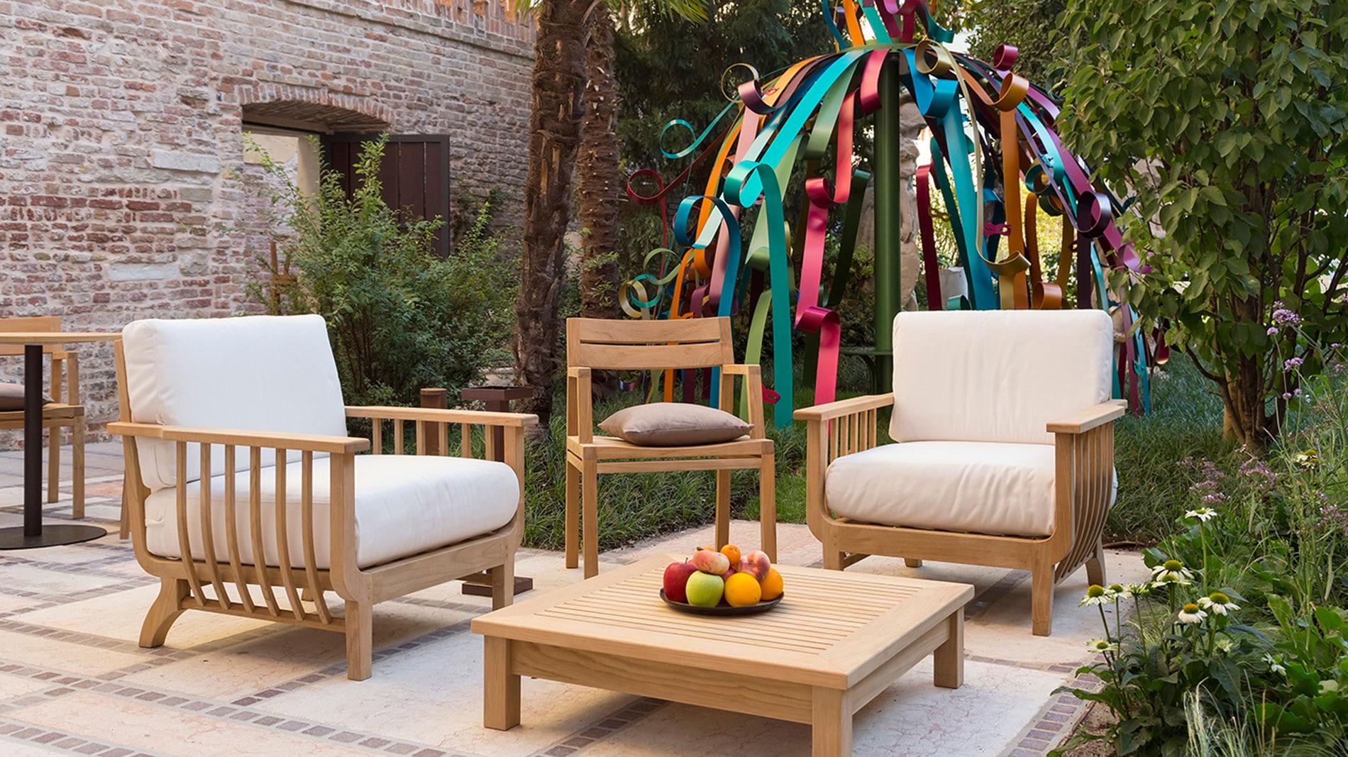 A garden seating area with wooden armchairs with cream cushions and a low table, next to a brick wall and a sculpture made of coloured ribbons.
