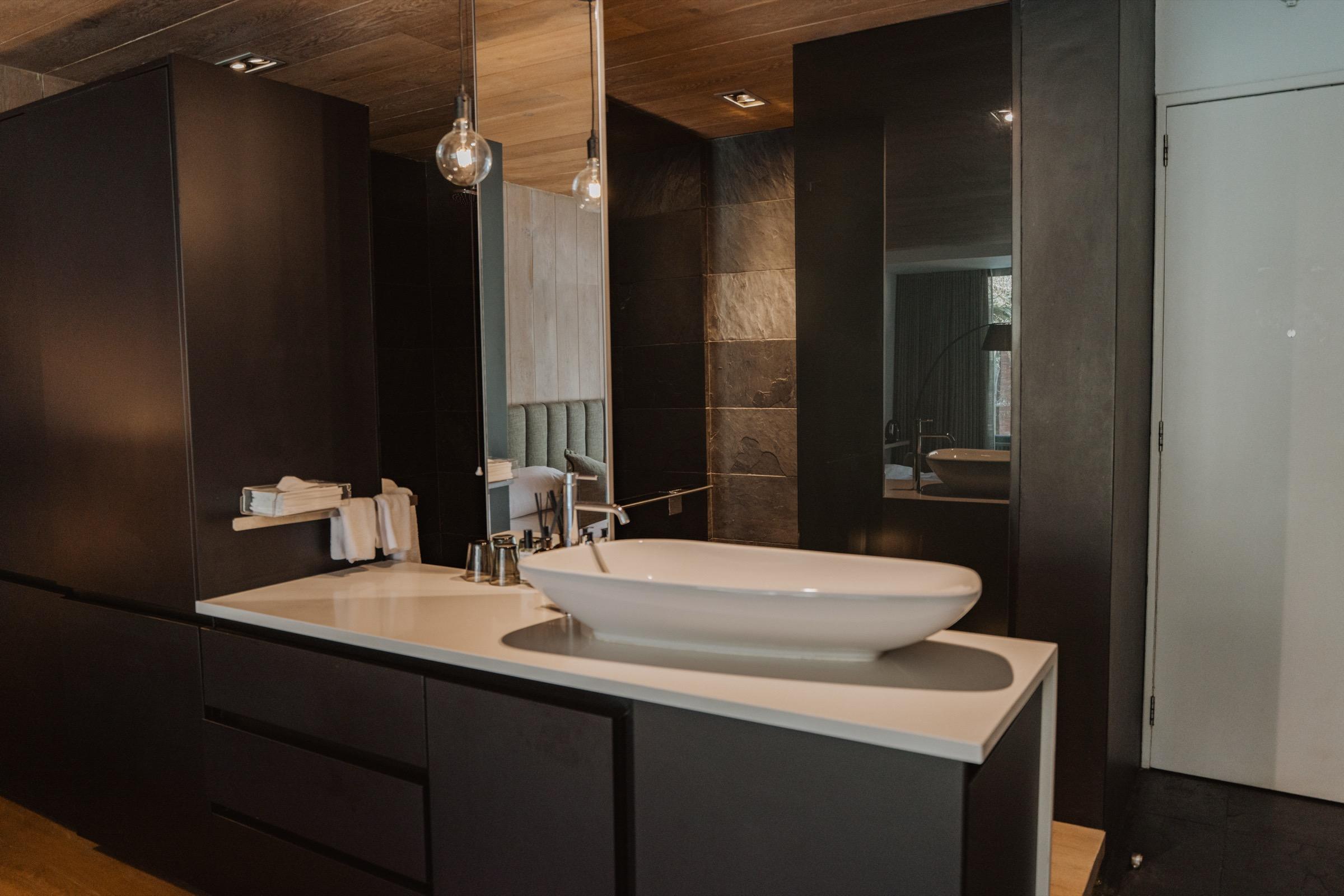 A black-cabinetry bathroom with an oval vessel basin, a wall-mounted gold pendant light and a mirror reflecting the bedroom beyond.