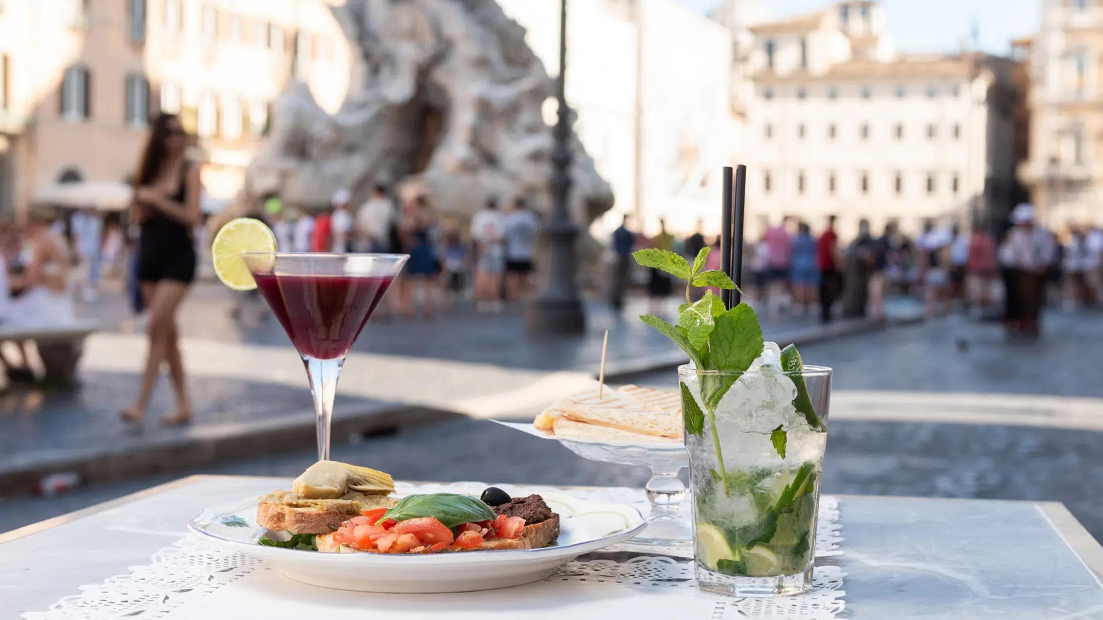 A drink and a plate on a Tre Scalini table, the fountains of Piazza Navona behind