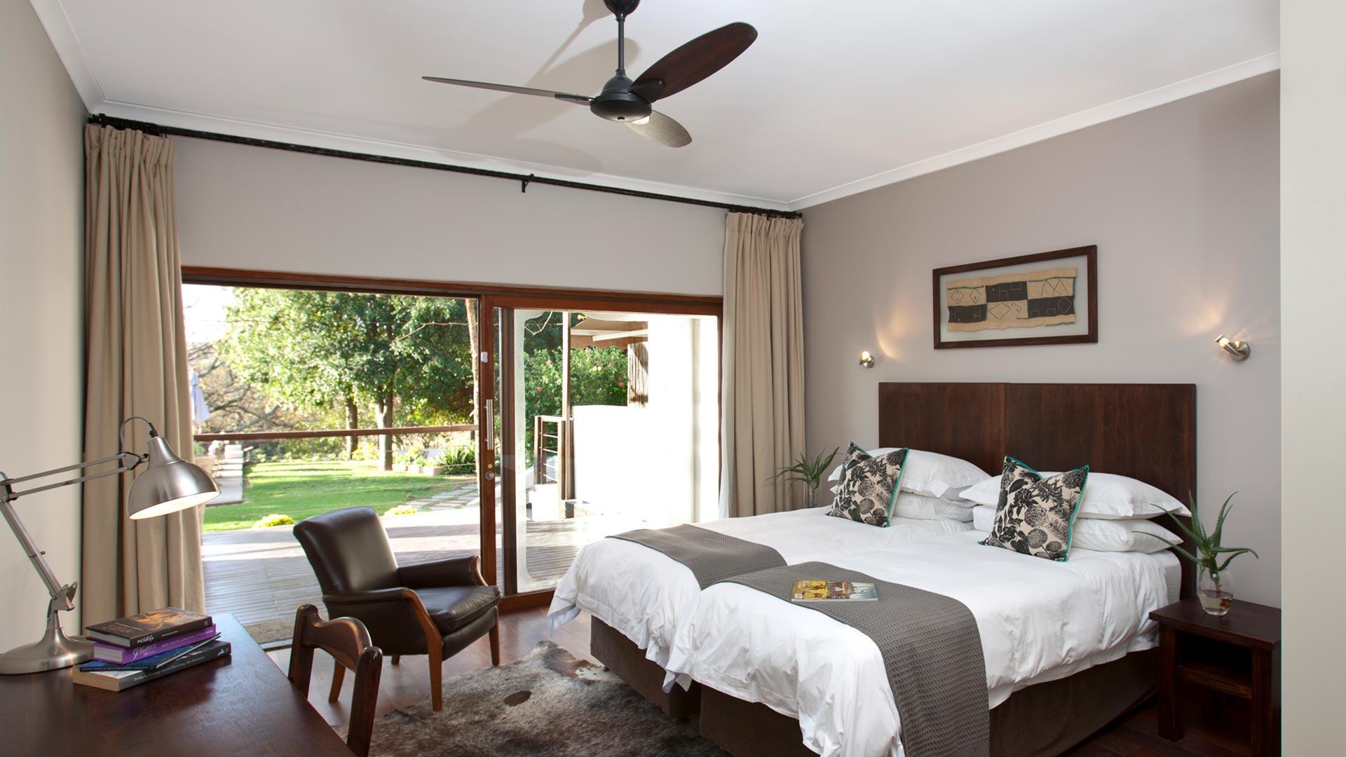 A bedroom with a king bed, a leather armchair and a desk lamp, with sliding glass doors on to a lawn and trees.