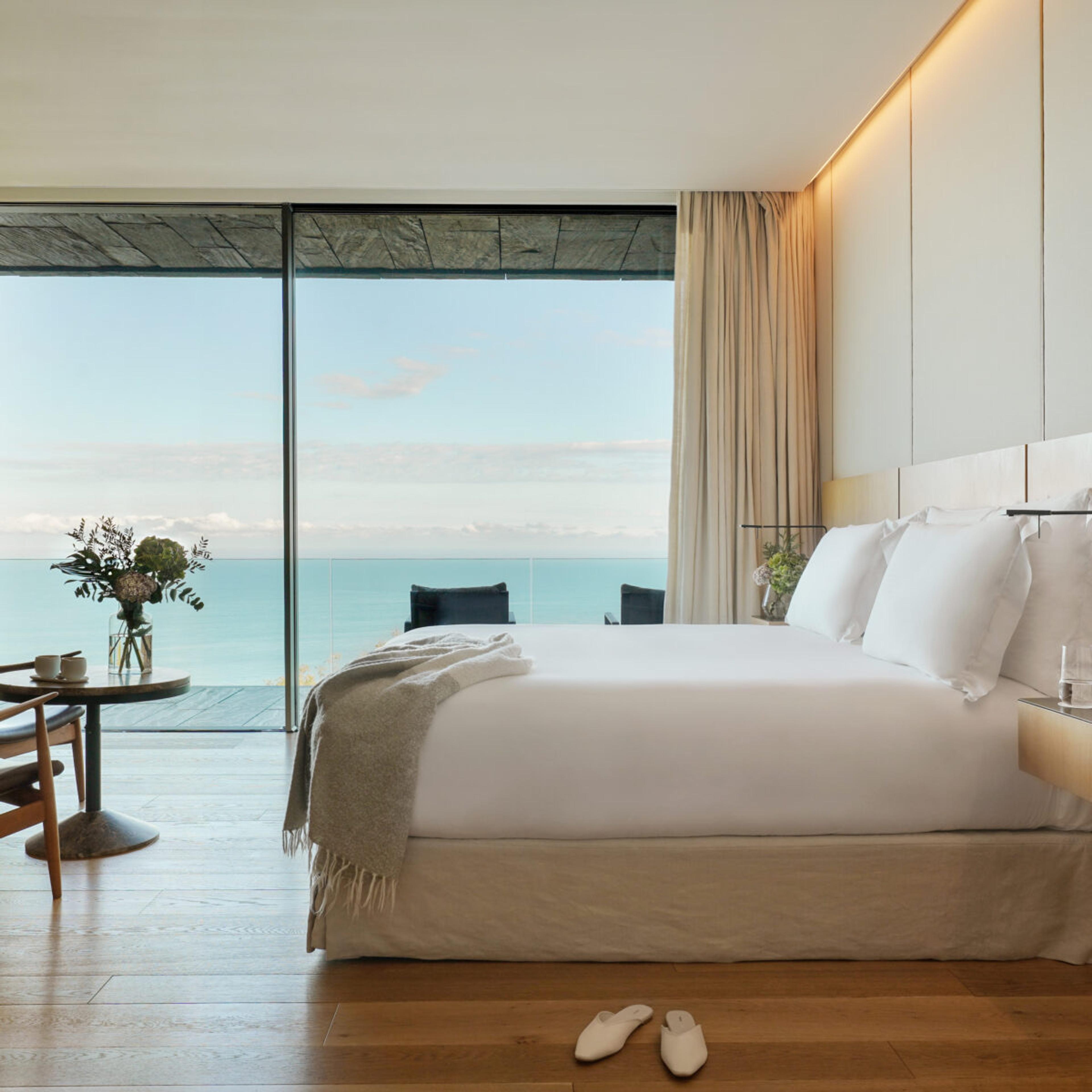 A bedroom bed dressed in white linen and a grey throw, with a round table set for two, fresh flowers, and a pair of white slippers on the floor, a glass door open to the sea.
