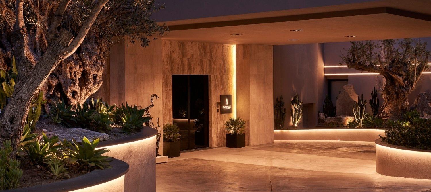 The hotel's entrance at night, lit stone walls either side of a glass door beneath olive trees.