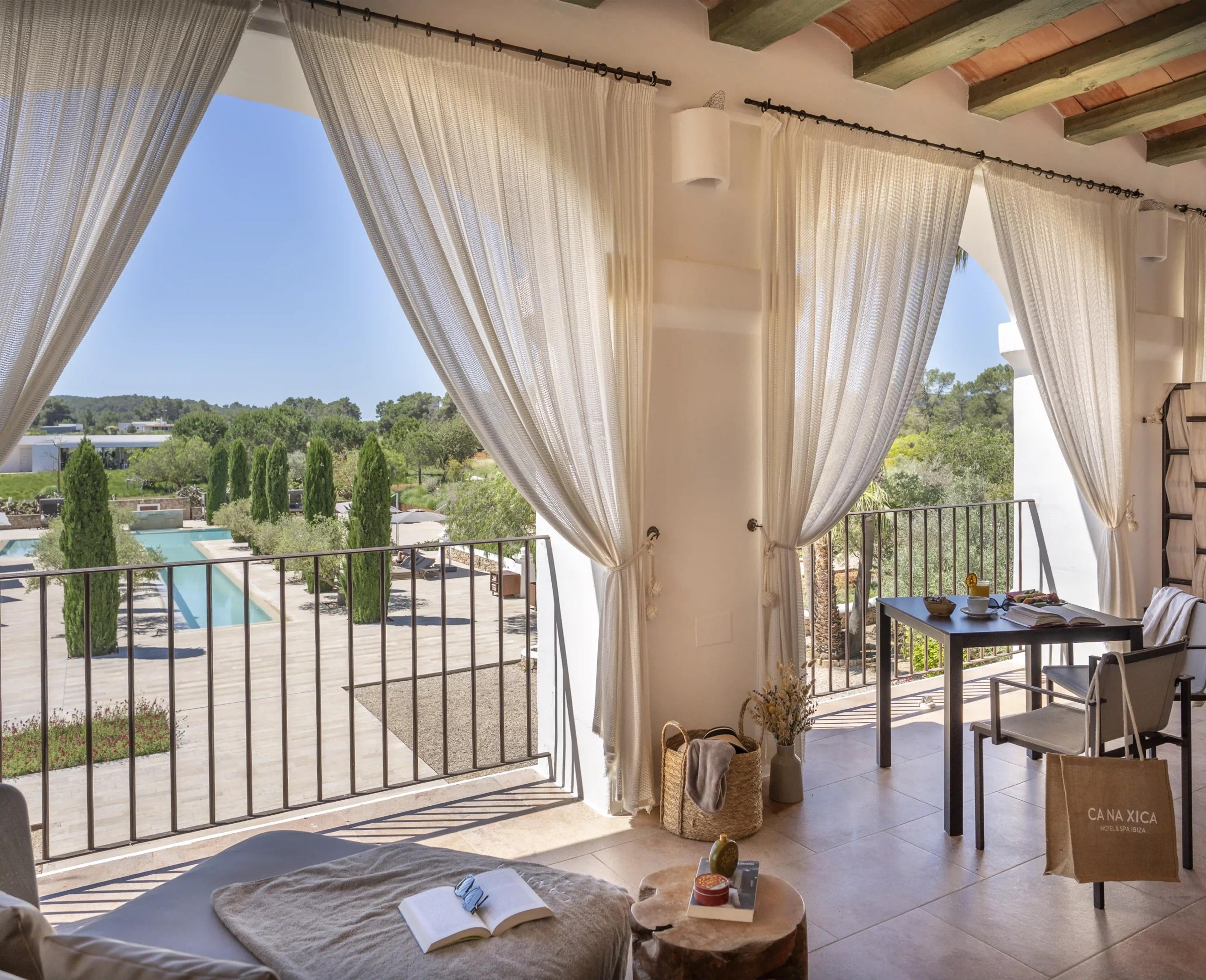 A covered terrace with sheer curtains drawn open on either side of a stone pillar, a table set with coffee and a book, and a swimming pool and cypress-lined garden below.