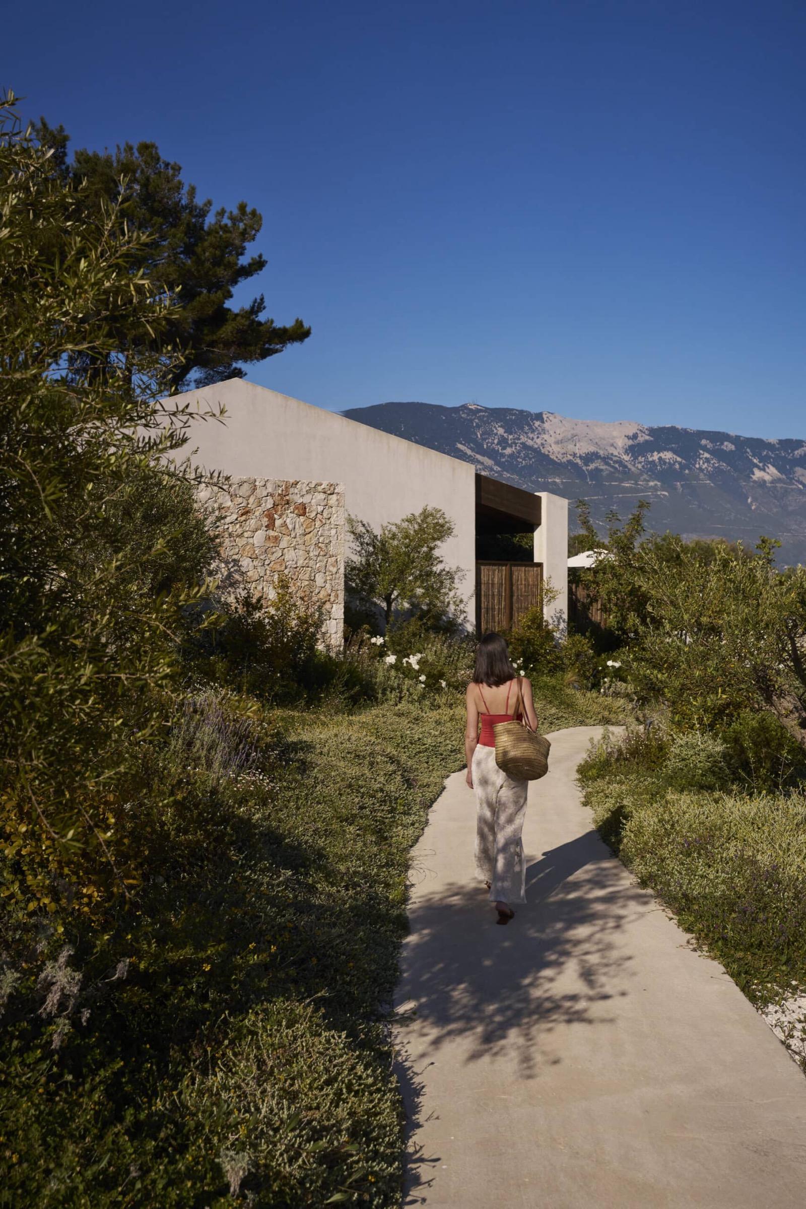 a woman in a white dress carrying a straw bag, walking away down a path towards a stone building, mountains behind her