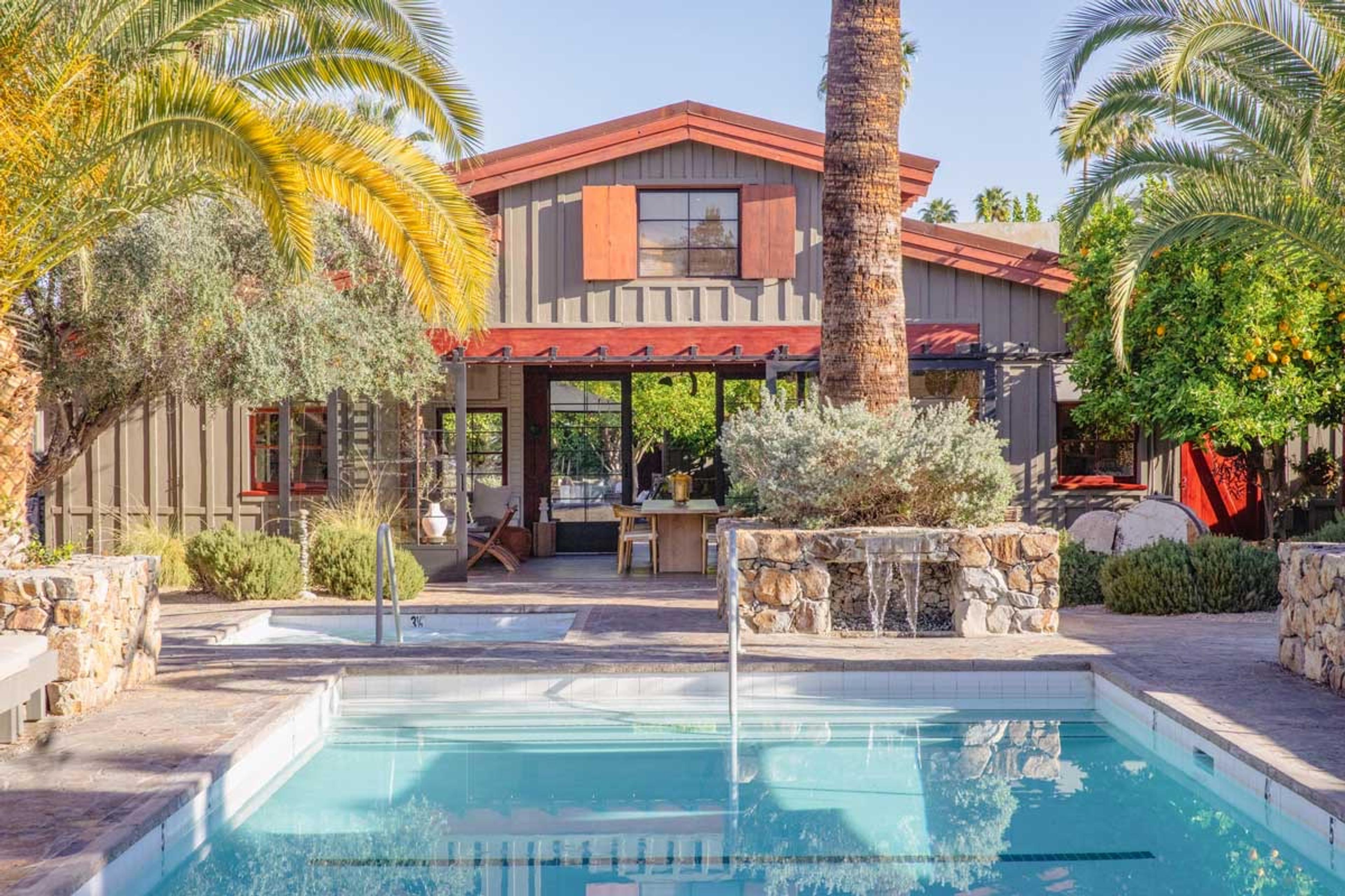 A swimming pool in daylight in front of a two-storey wooden house with a red-trimmed roof, with palm trees either side and a stone planter with a small fountain.
