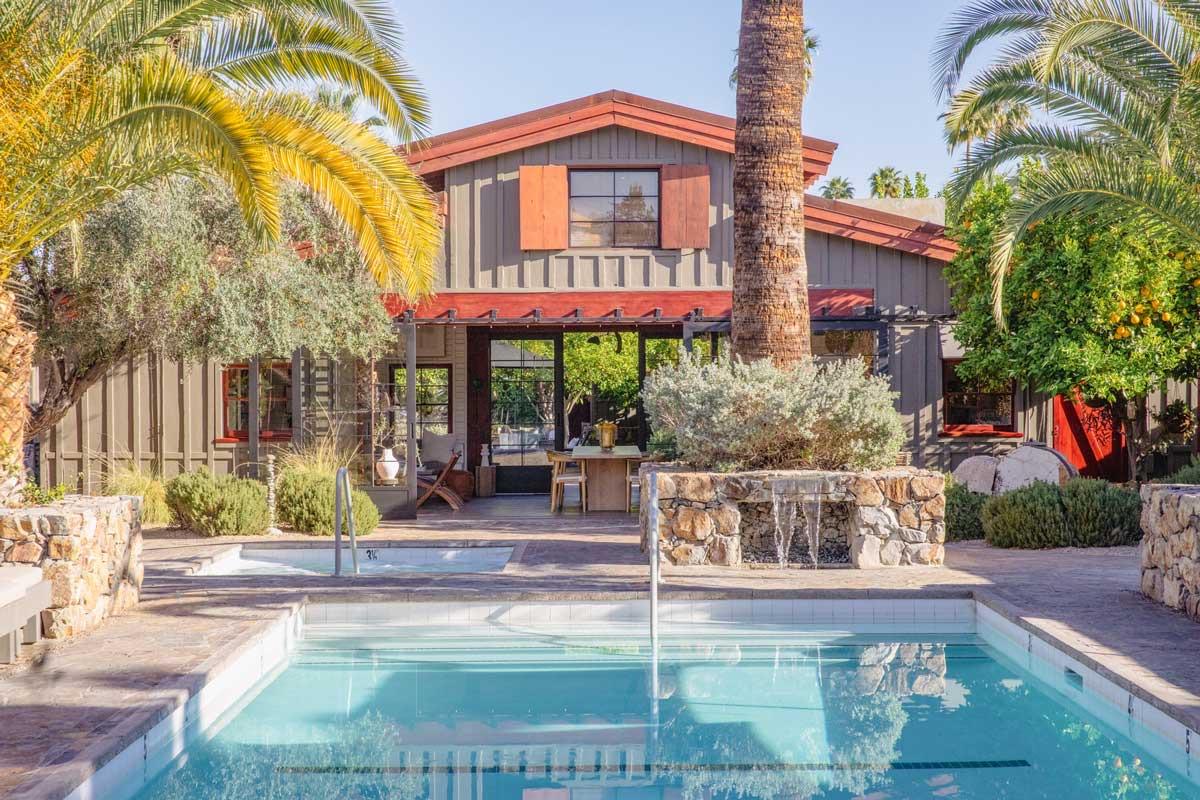 A swimming pool in daylight in front of a two-storey wooden house with a red-trimmed roof, with palm trees either side and a stone planter with a small fountain.