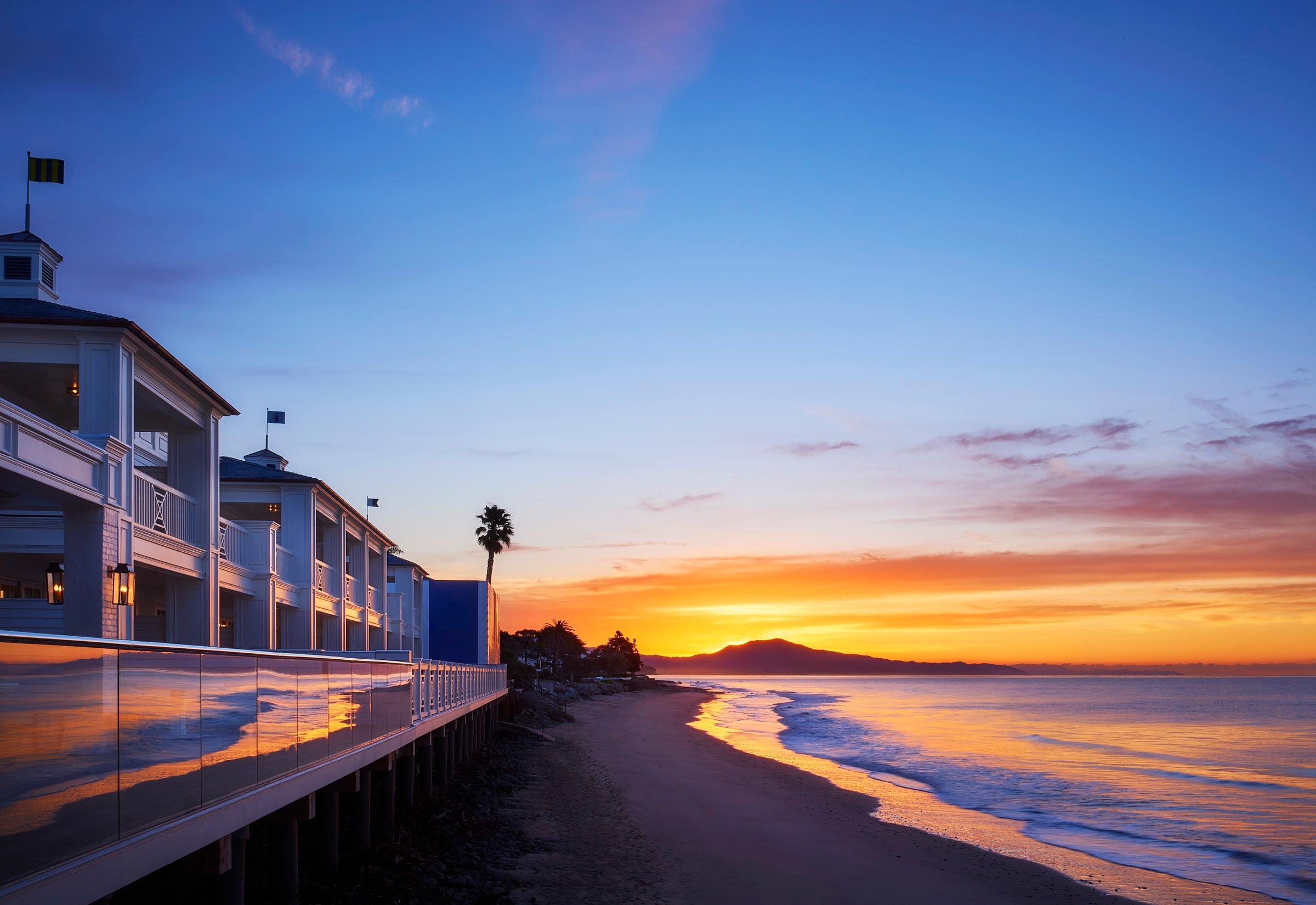 A row of white, shingled buildings on pilings along a beach at sunset, with a palm tree and mountains across the water.