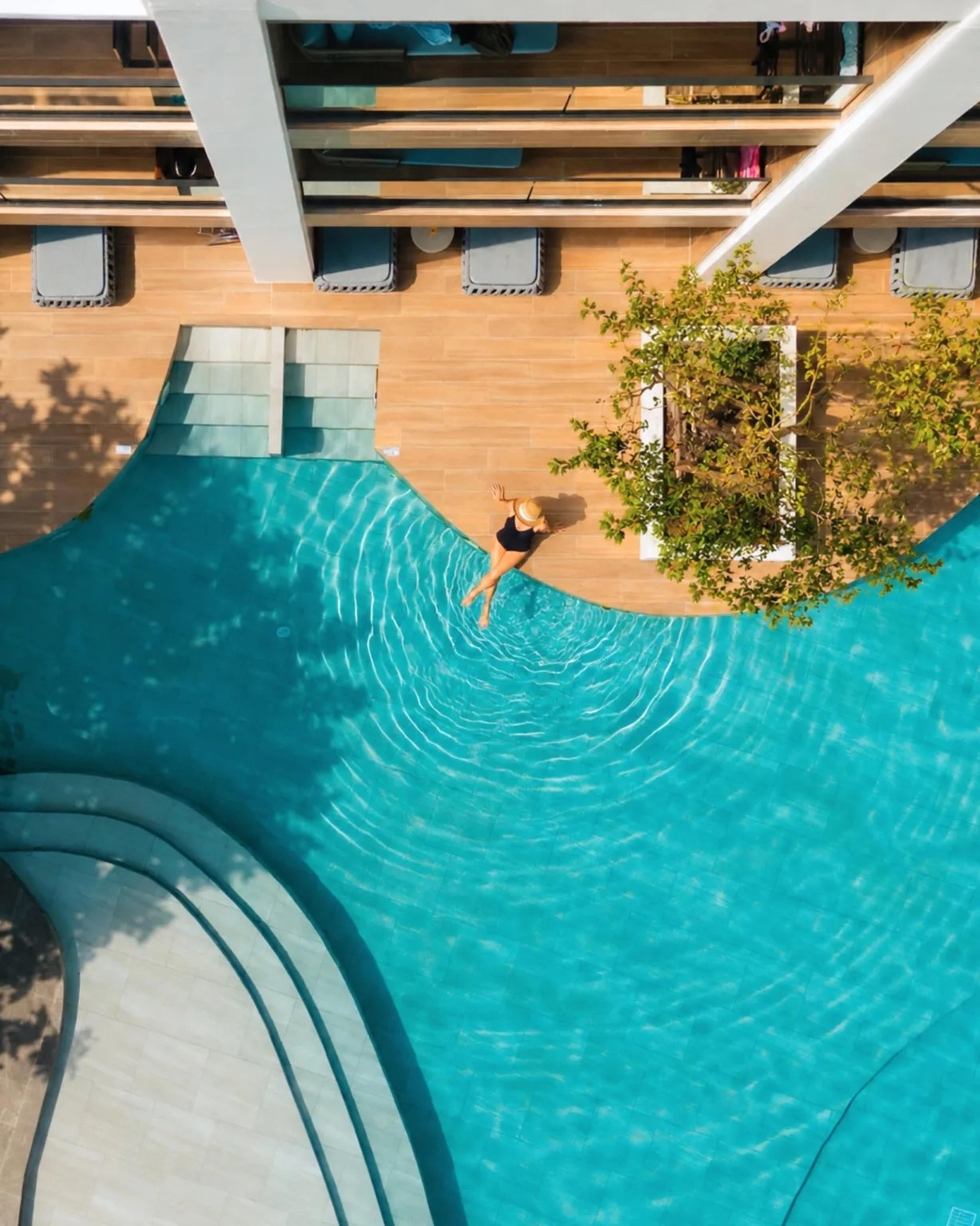an aerial view of a woman in a sunhat wading into a curved swimming pool, seen from directly above