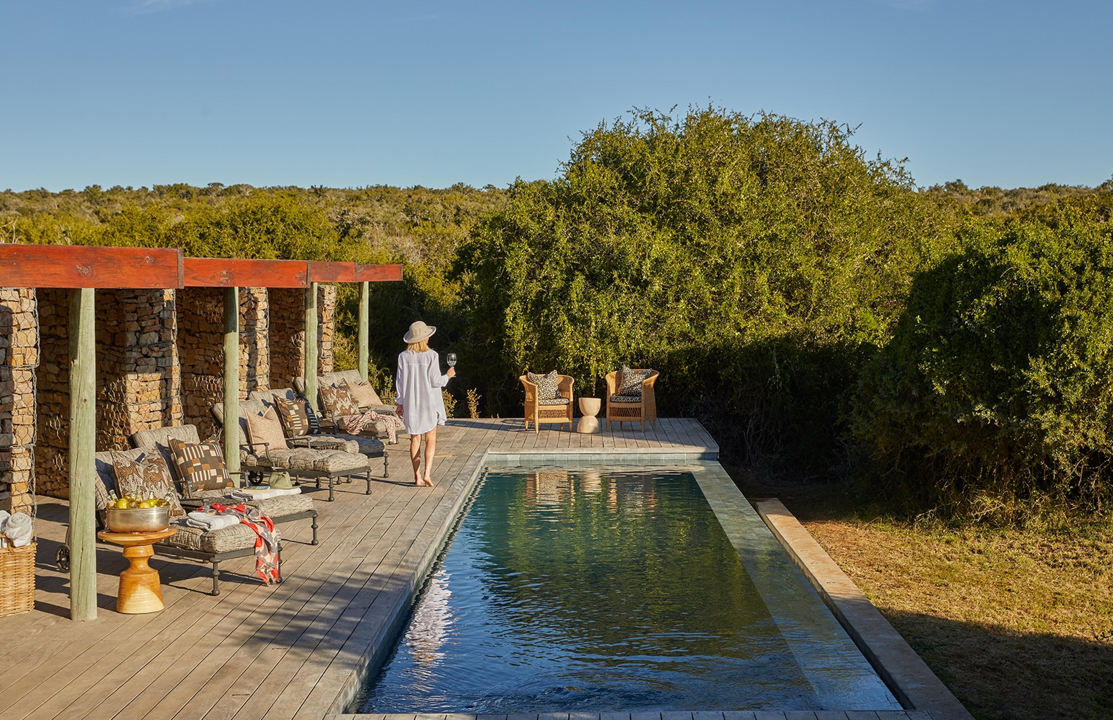A woman in a sunhat walking along a pool deck past loungers under a stone colonnade, with bush beyond.