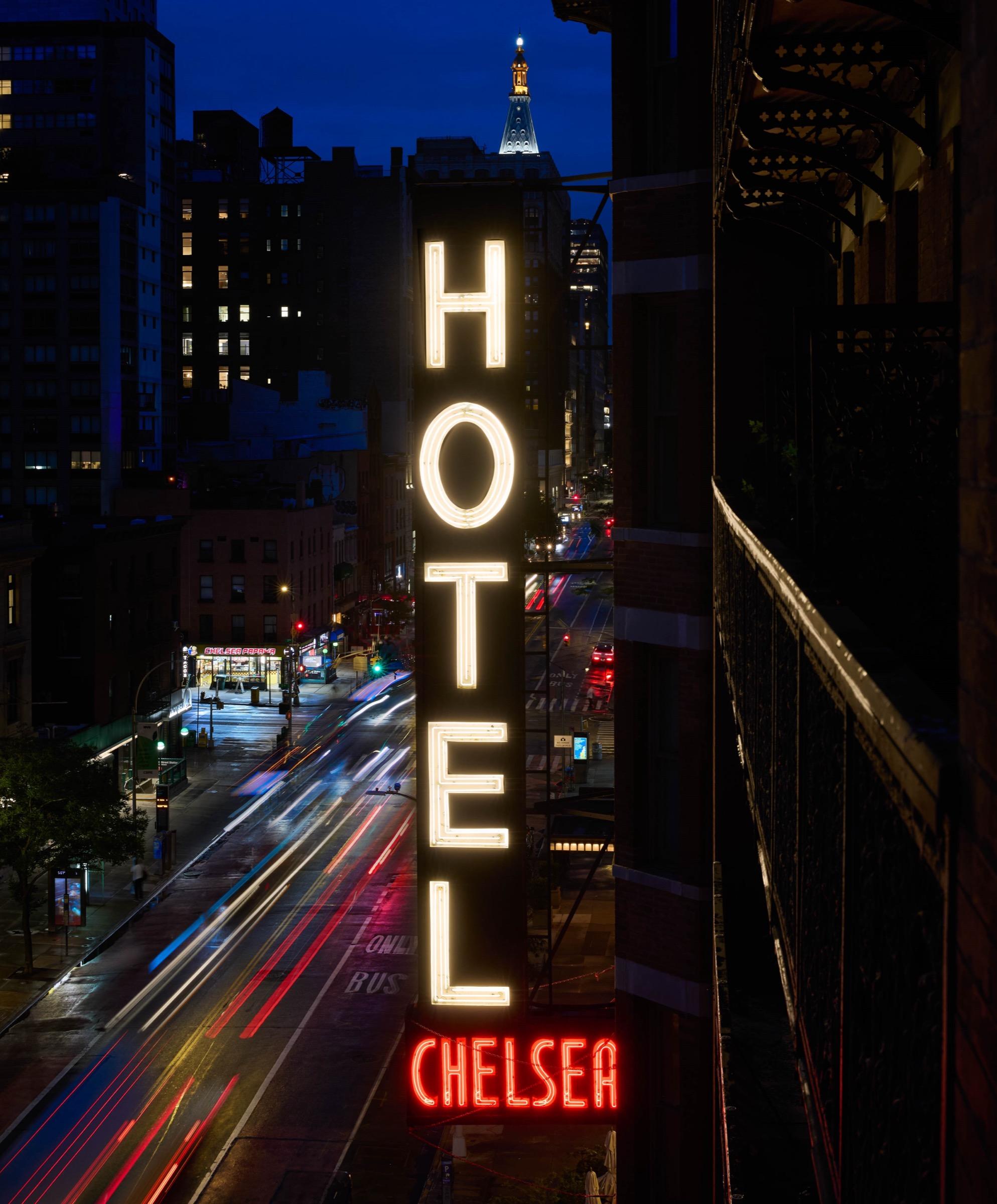 A vertical Hotel Chelsea sign glows above a city street at night, with light trails from passing traffic and a distant illuminated spire.
