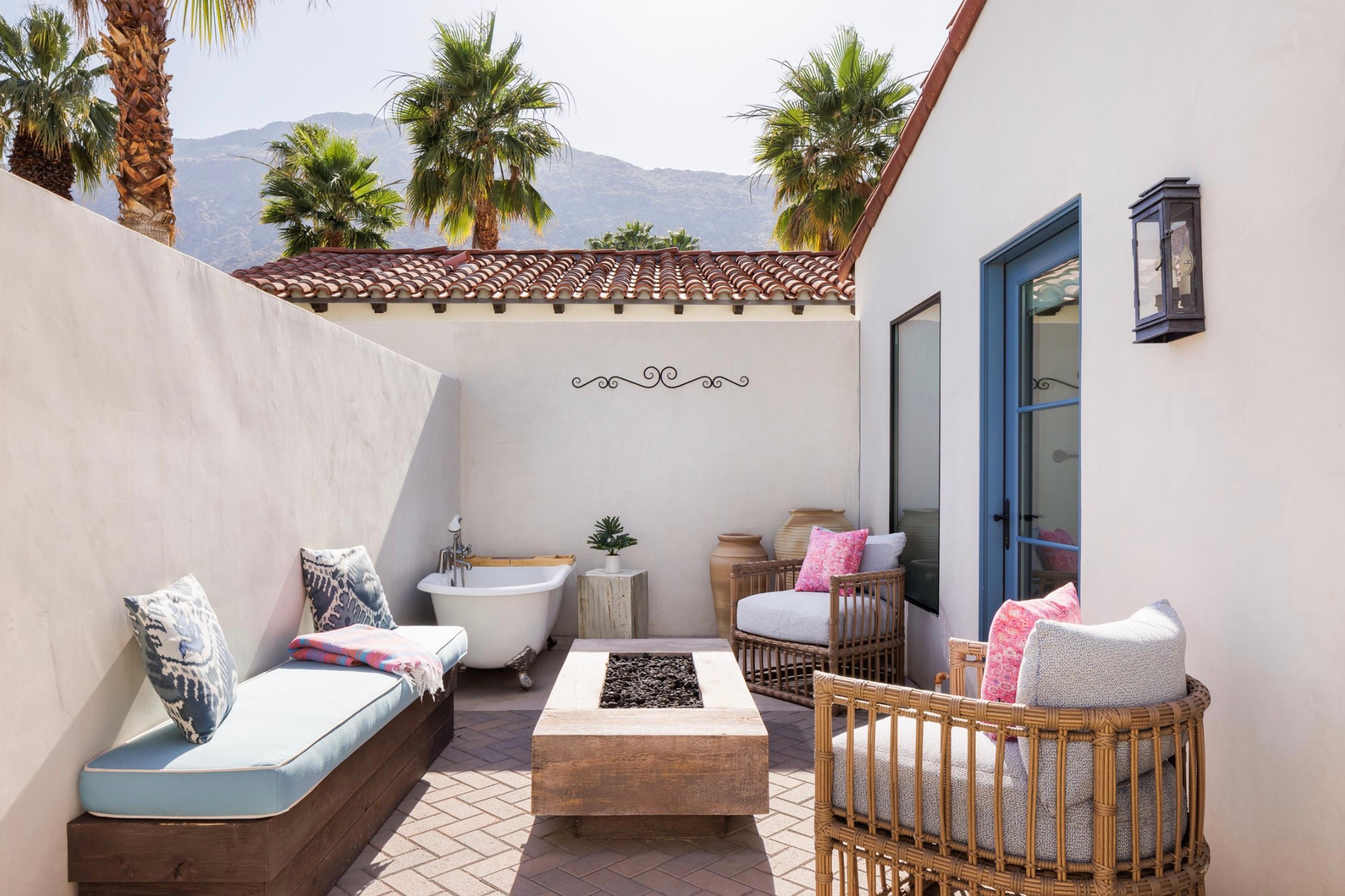 A private walled patio with a built-in bench, a stone fire pit, a freestanding bathtub and rattan armchairs, palm trees and mountains behind.