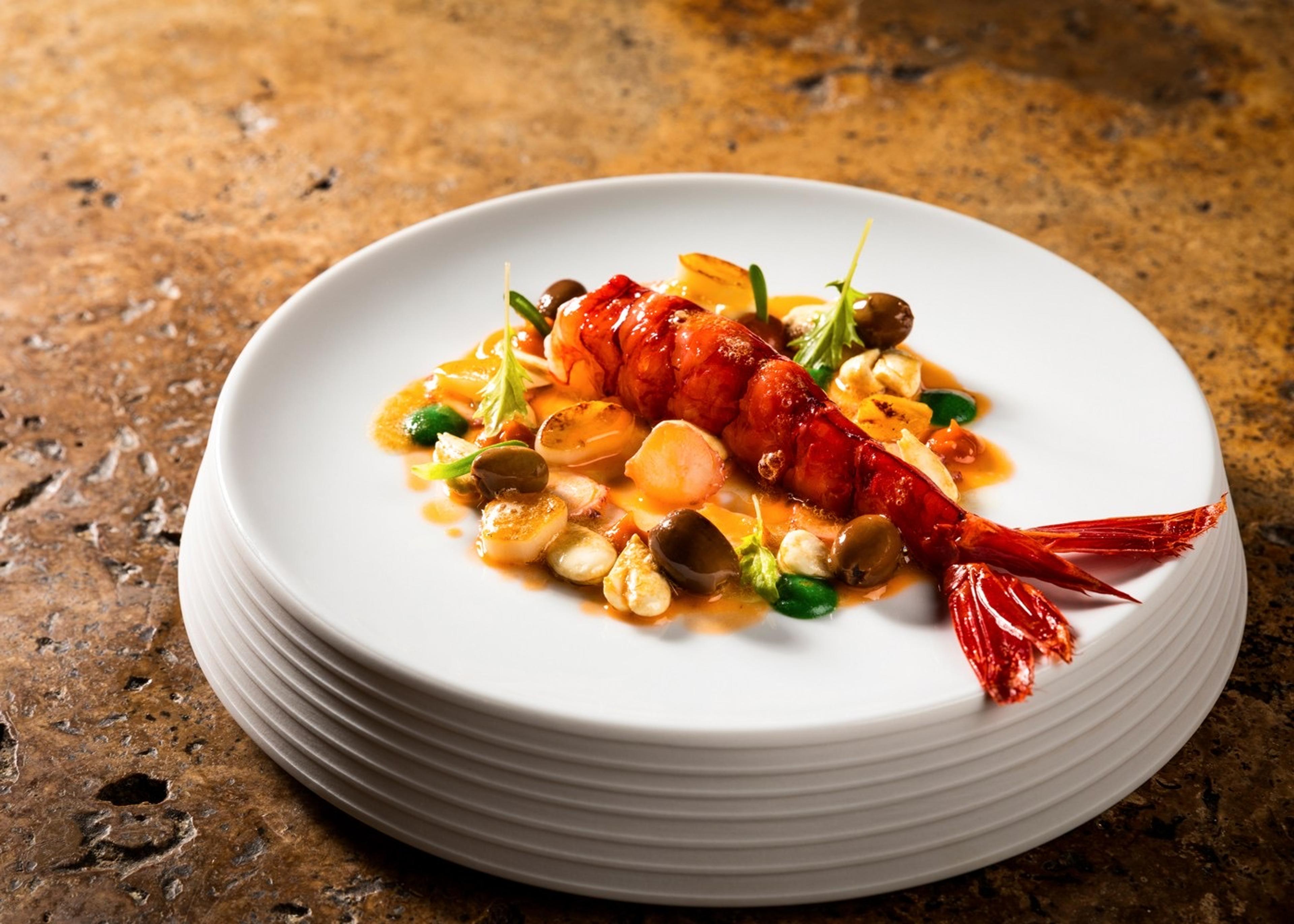 A whole grilled langoustine plated with shellfish, olives and herbs in a pool of sauce on a white plate.
