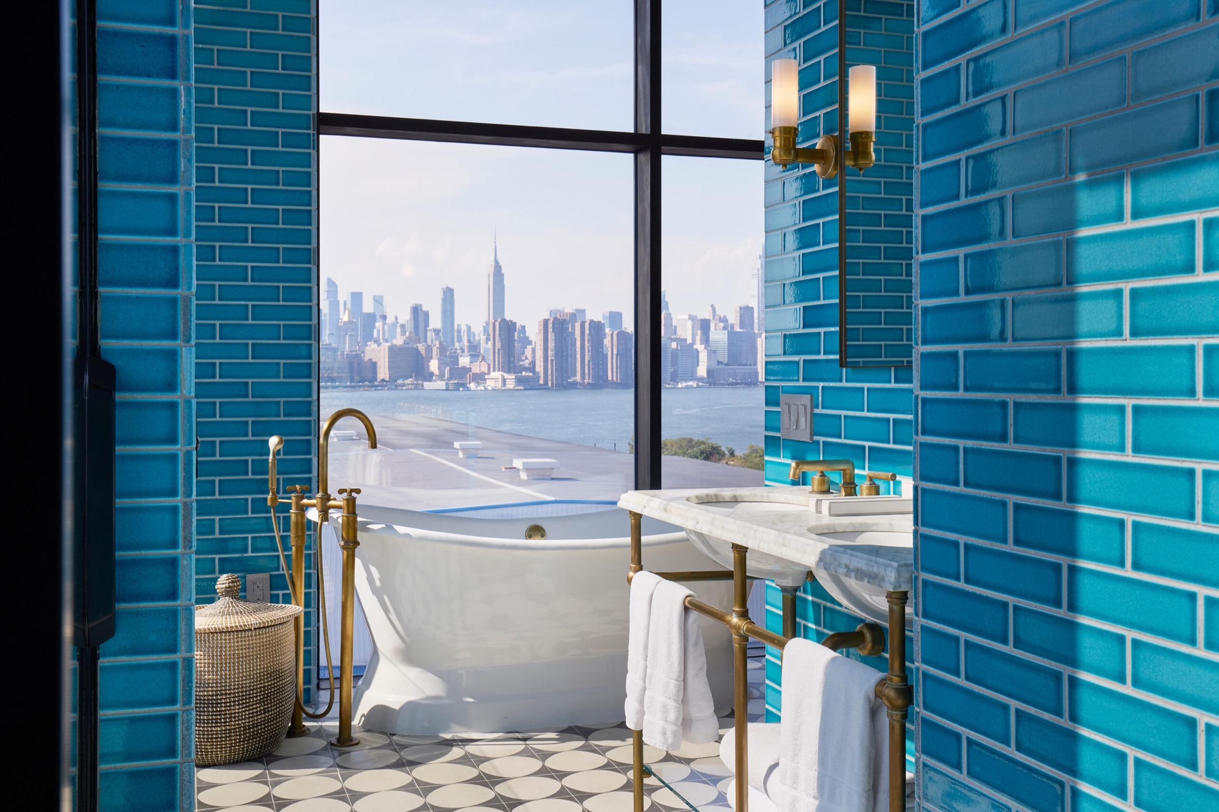 A freestanding white soaking tub and marble sink in a teal tiled bathroom, with a floor-to-ceiling window framing the Manhattan skyline and river.