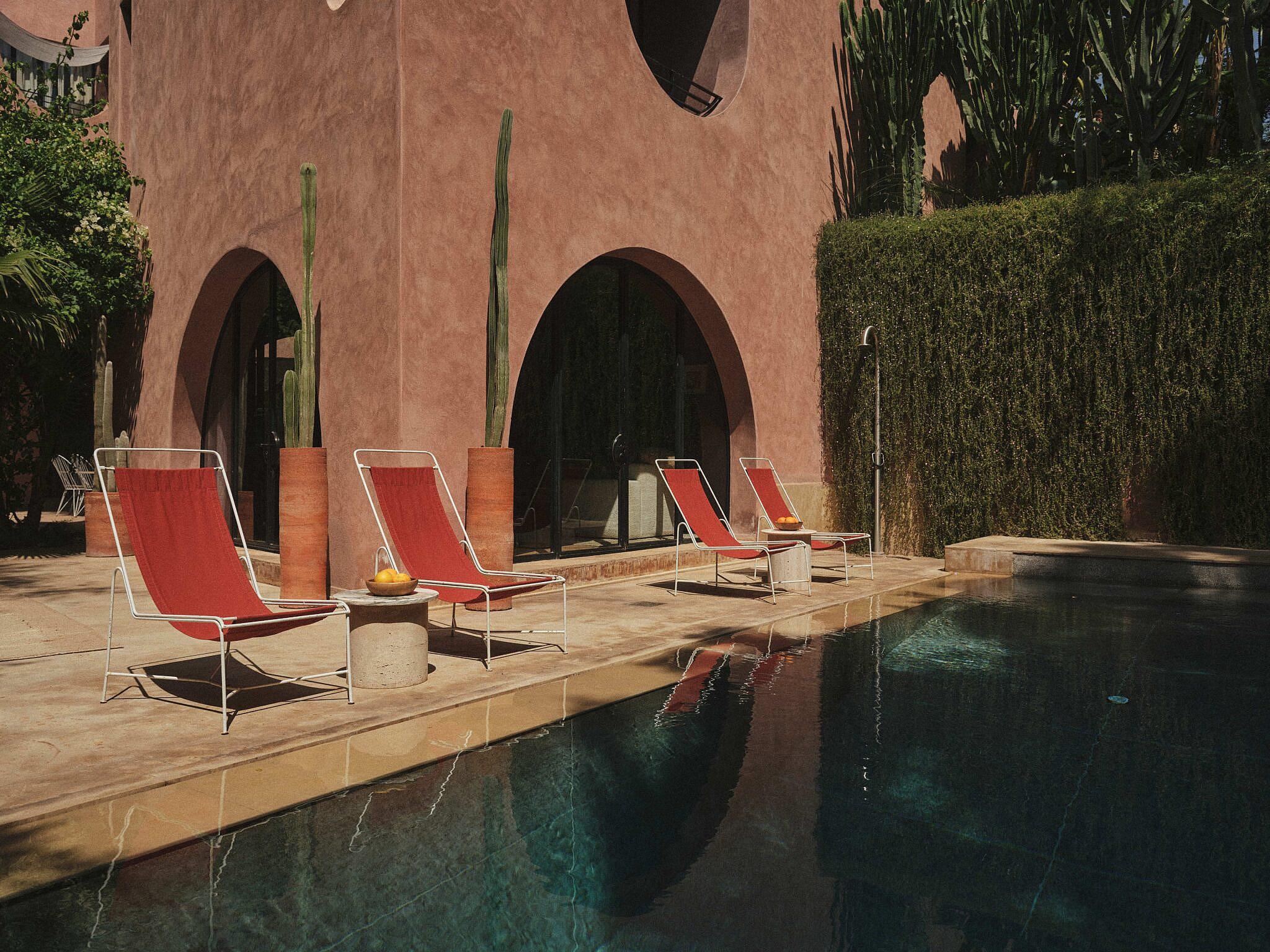 a pink stuccoed building with two arched doorways, four red lounge chairs in pairs on a terrace beside a pool, a wall of trailing vines alongside