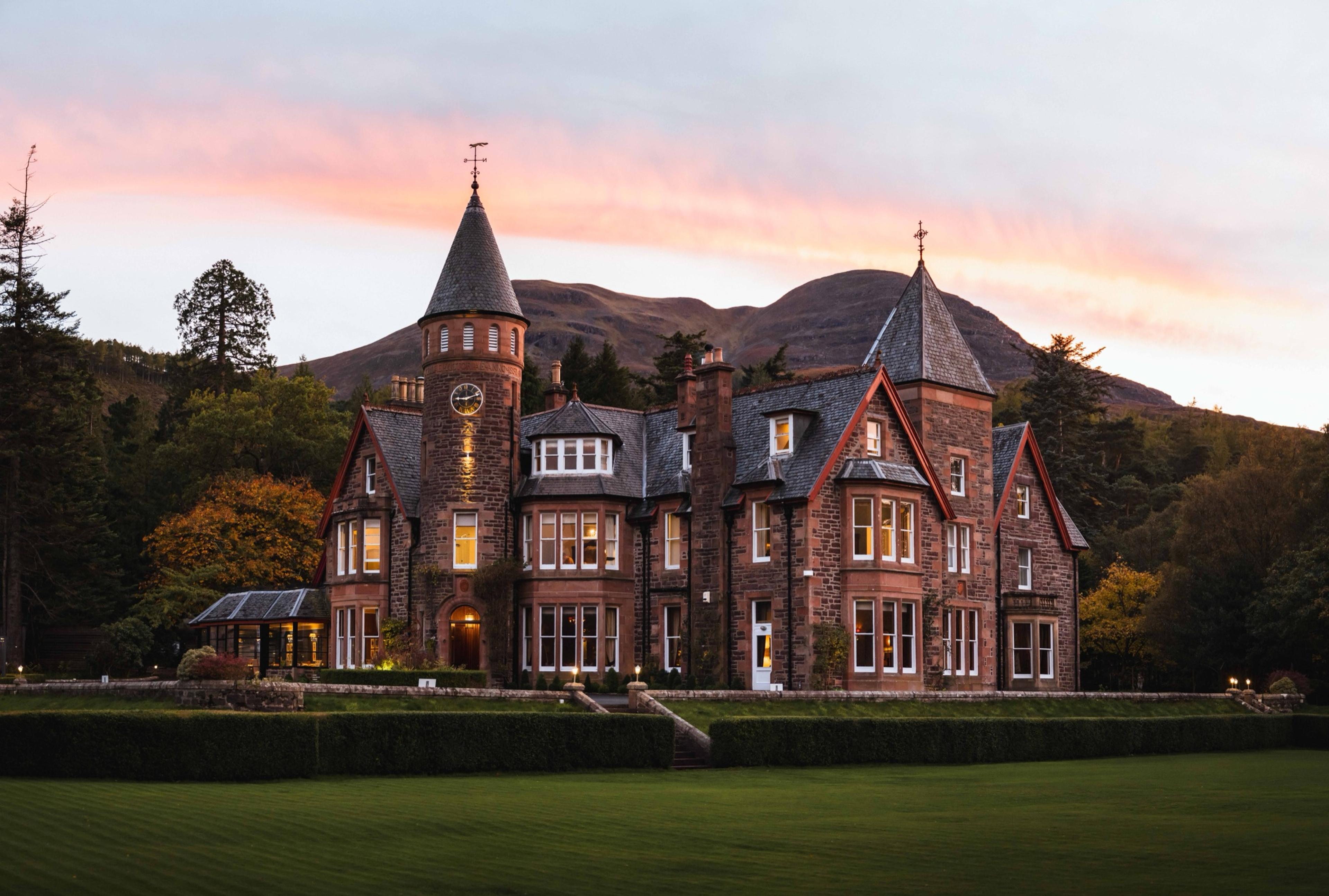 A turreted stone country house at dusk, its windows lit, set against hills under a pink and orange sky.