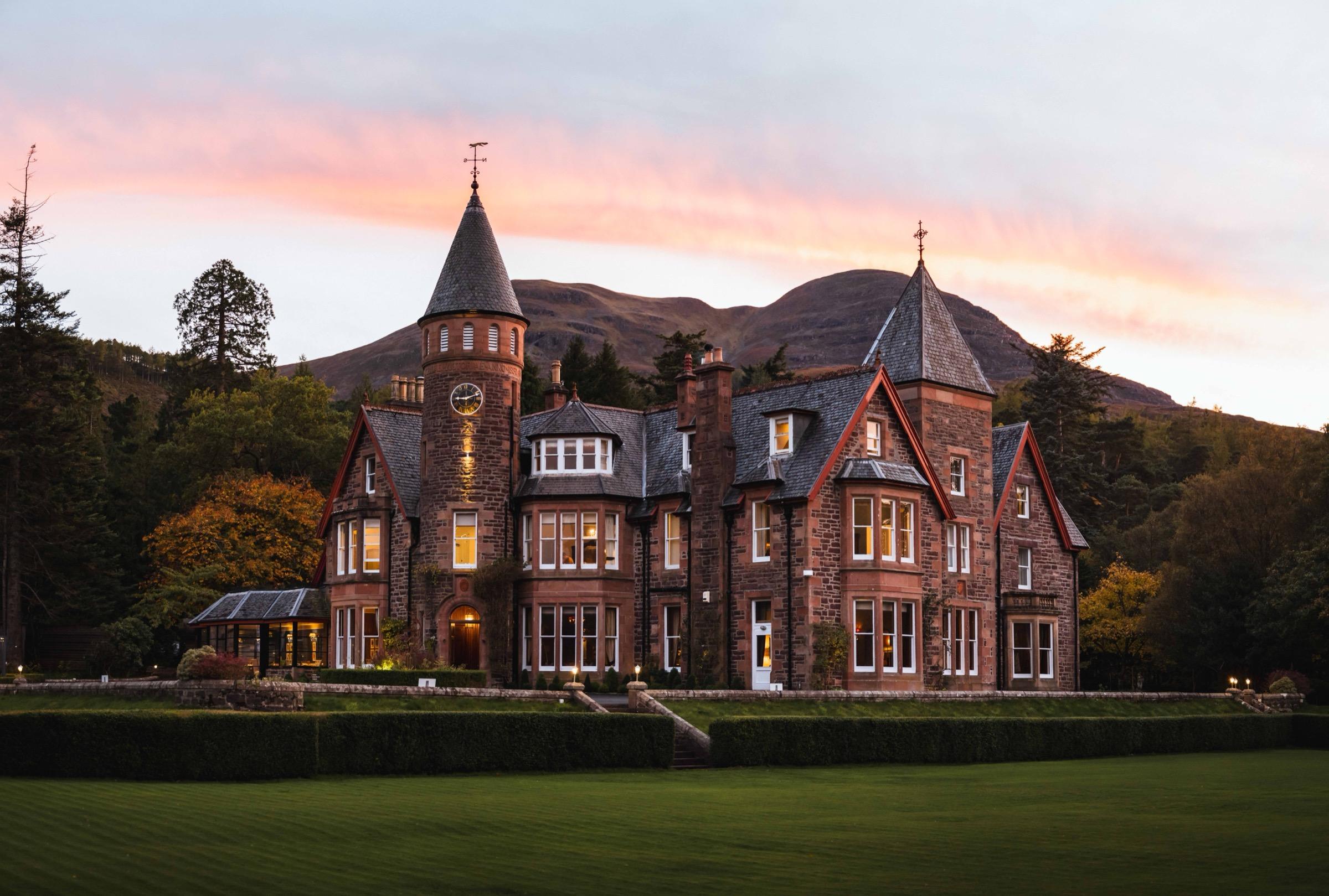 A turreted stone country house at dusk, its windows lit, set against hills under a pink and orange sky.