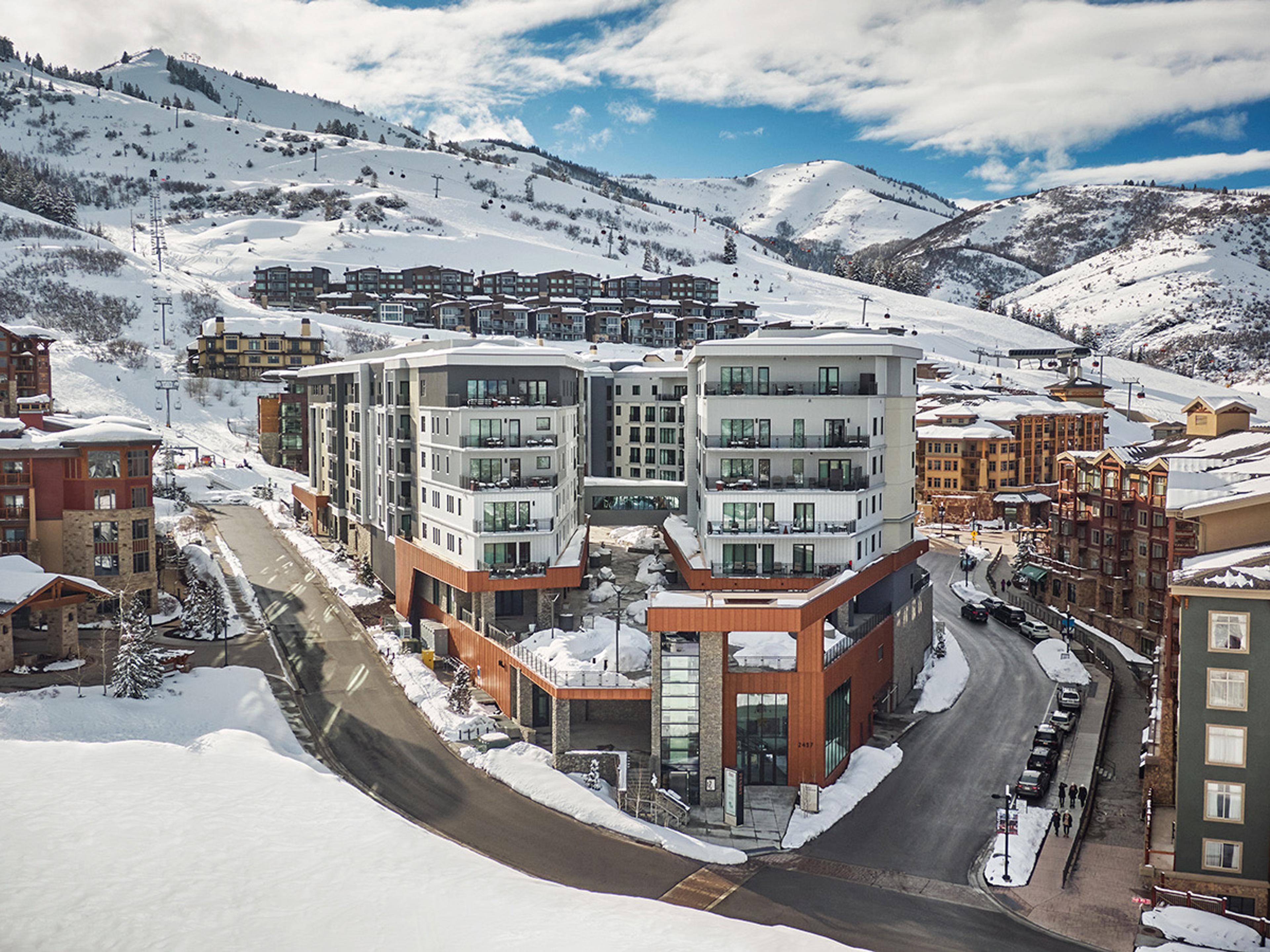 Aerial view of a multi-storey hotel building with white panels, balconies and orange wood cladding, set on a snowy hillside beside a curving road, with forested mountains behind under a blue sky with cloud.
