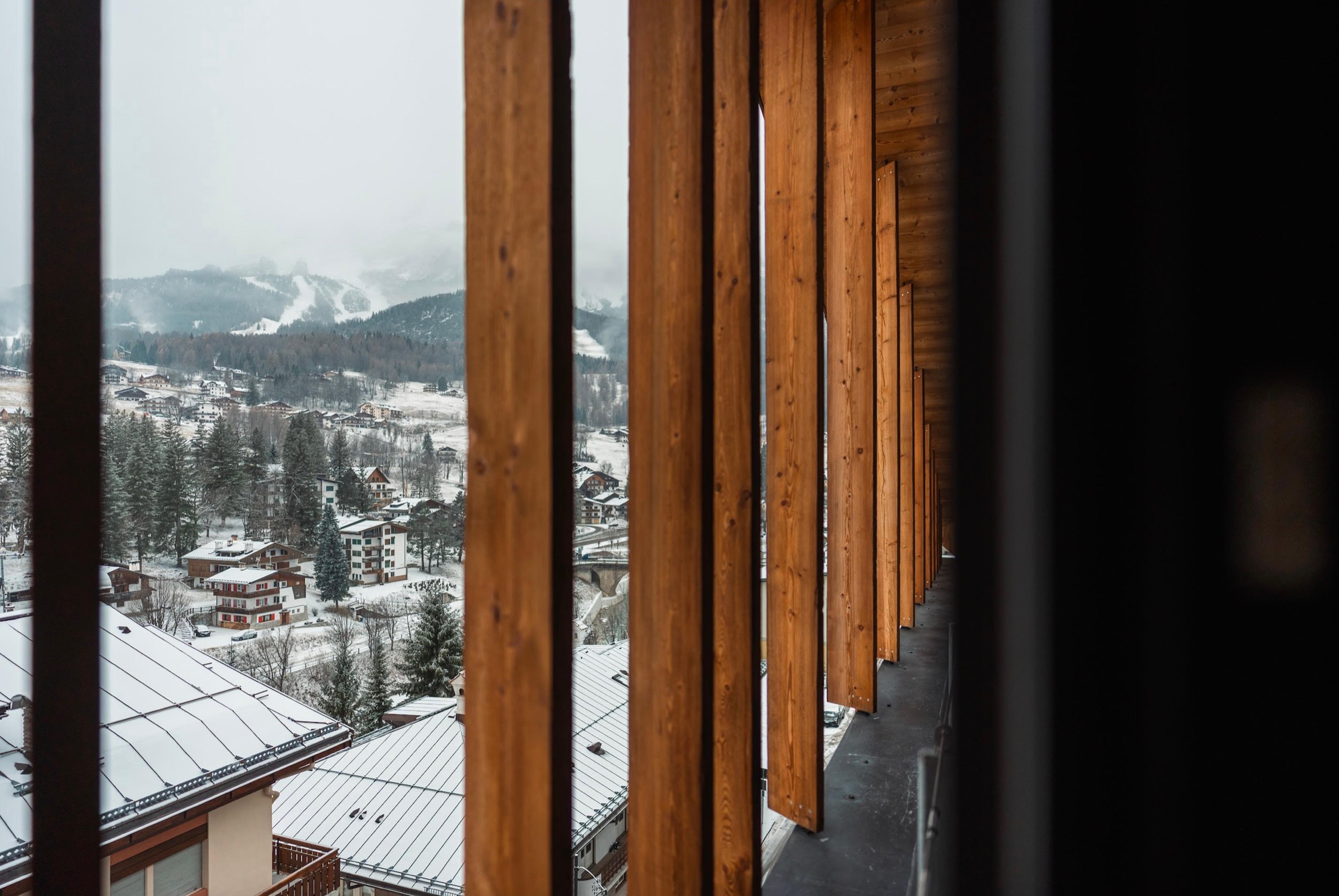 Snow-covered rooftops and pine trees are seen through a row of vertical timber slats on a balcony.