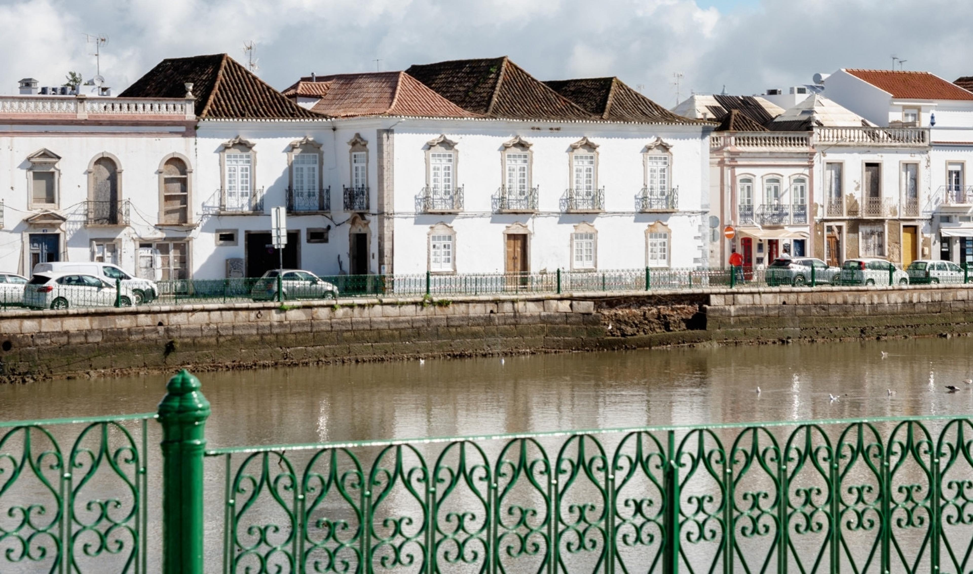 Whitewashed townhouses with tiled roofs along a riverbank, parked cars and a green wrought iron railing in the foreground, reflected in the water.
