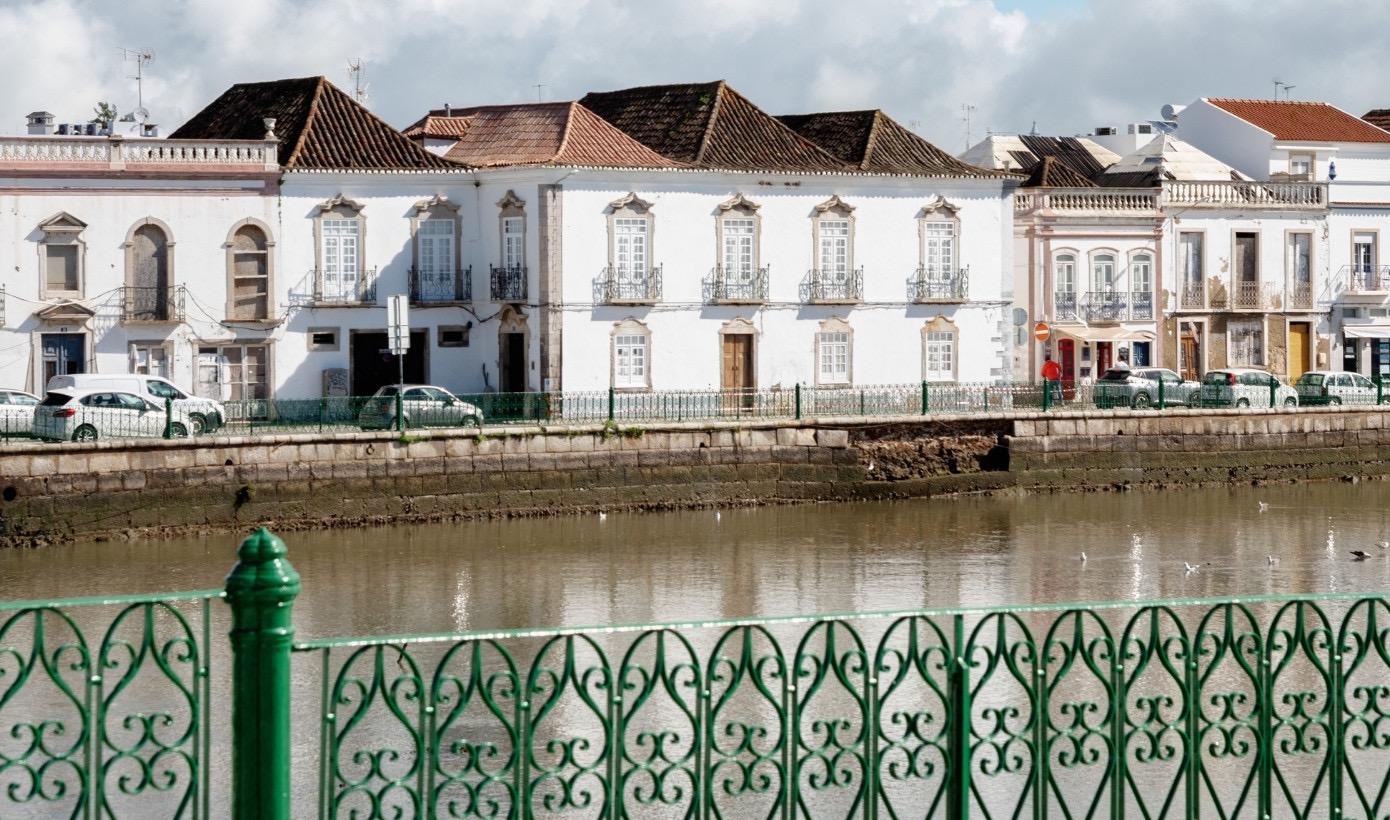 Whitewashed townhouses with tiled roofs along a riverbank, parked cars and a green wrought iron railing in the foreground, reflected in the water.