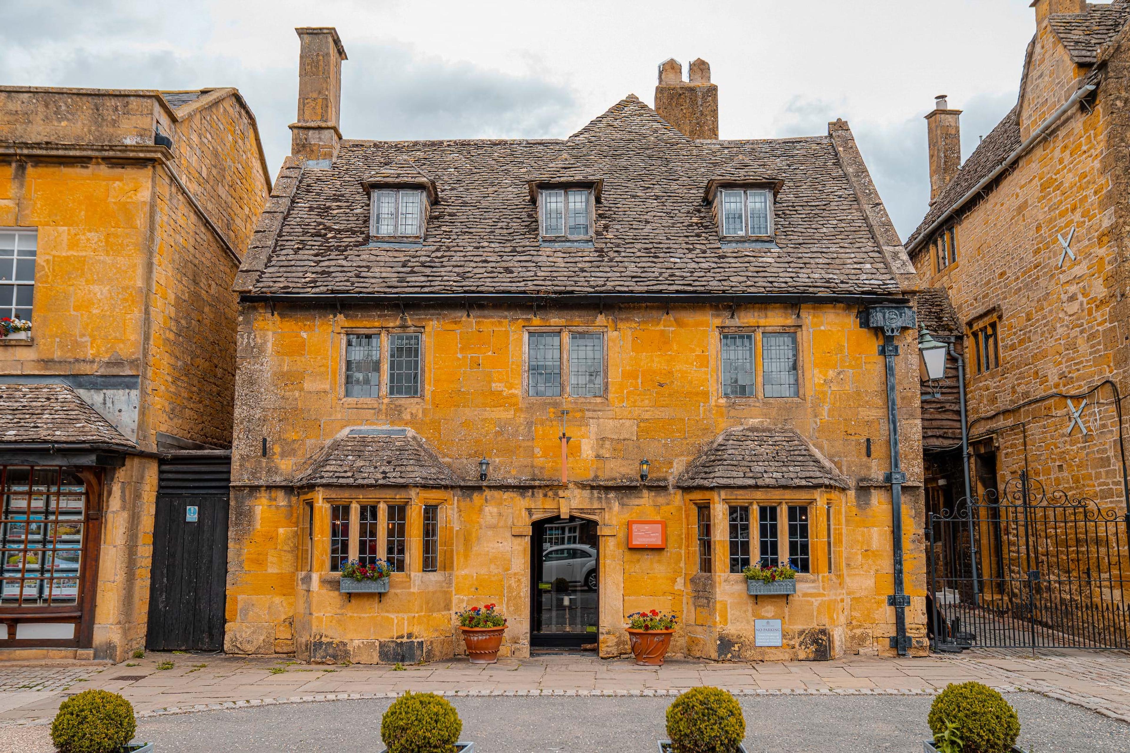 A three-storey stone building with dormer windows, leaded windows, and potted flowers either side of its arched doorway.