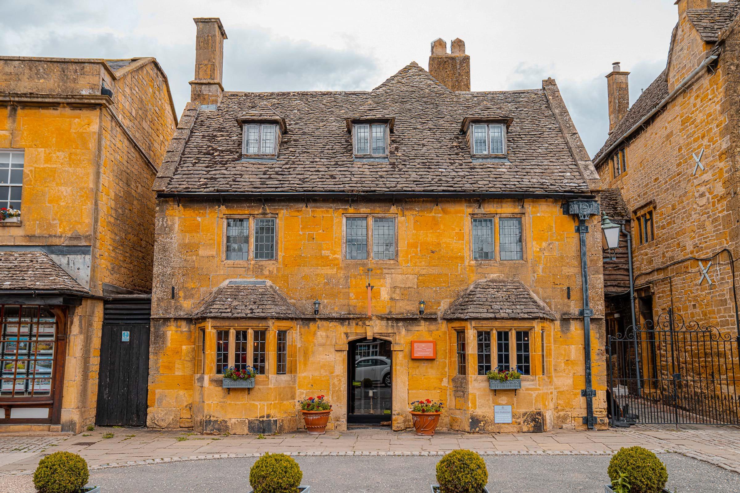 A three-storey stone building with dormer windows, leaded windows, and potted flowers either side of its arched doorway.
