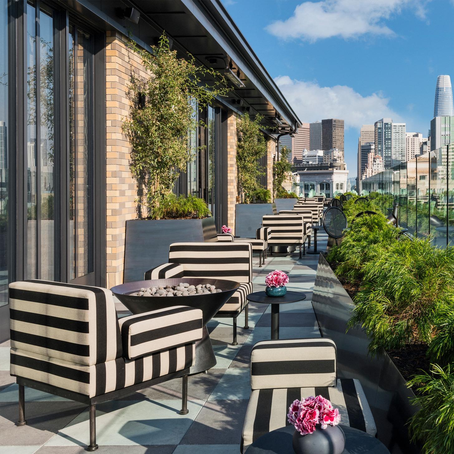 A daytime rooftop terrace with black and white striped chairs, potted greenery along a glass railing, and the San Francisco skyline including a tall glass tower.