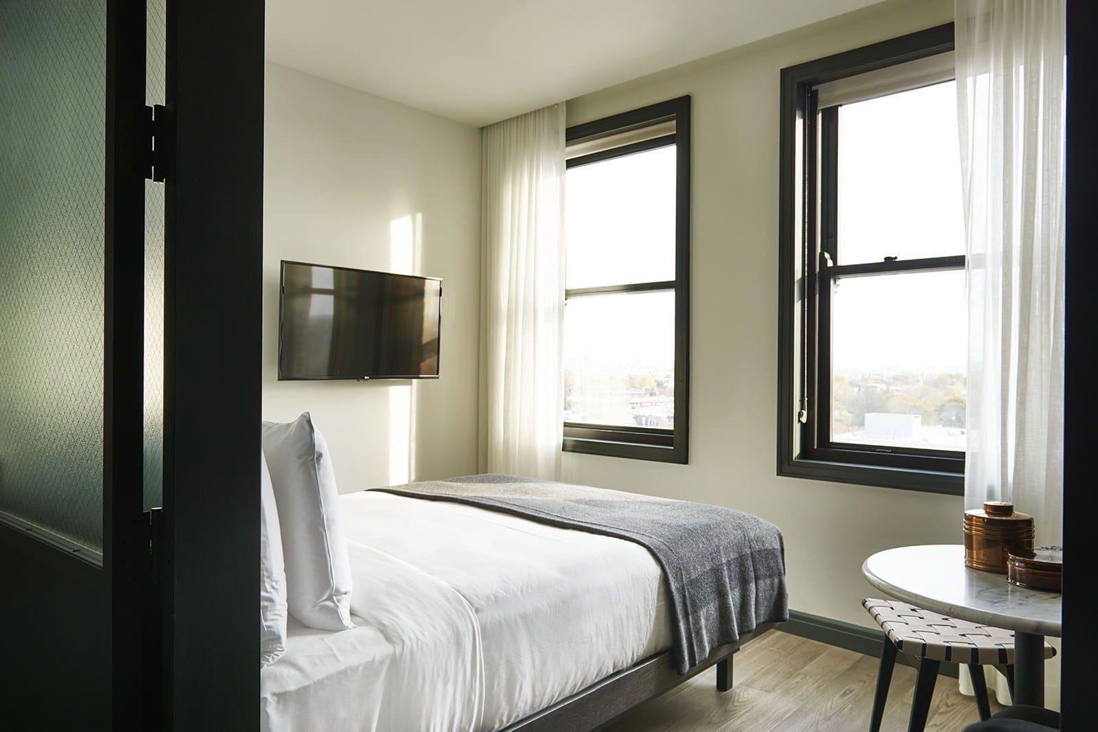 a bed with a folded grey blanket on white linens, facing two tall black-framed windows with sheer curtains showing a hazy city skyline, a small round table and woven stool to the right