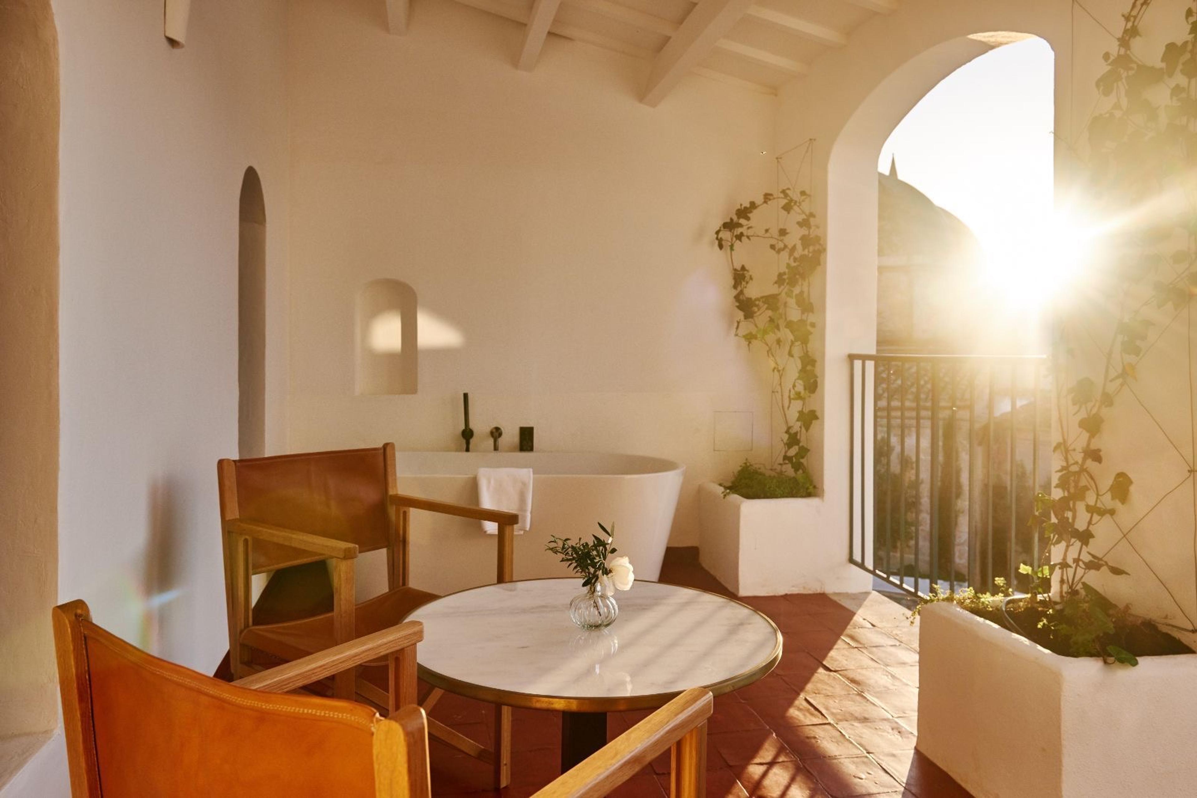 A freestanding bathtub and two leather chairs around a marble table on a covered terrace, with low sun flaring through an arched opening.