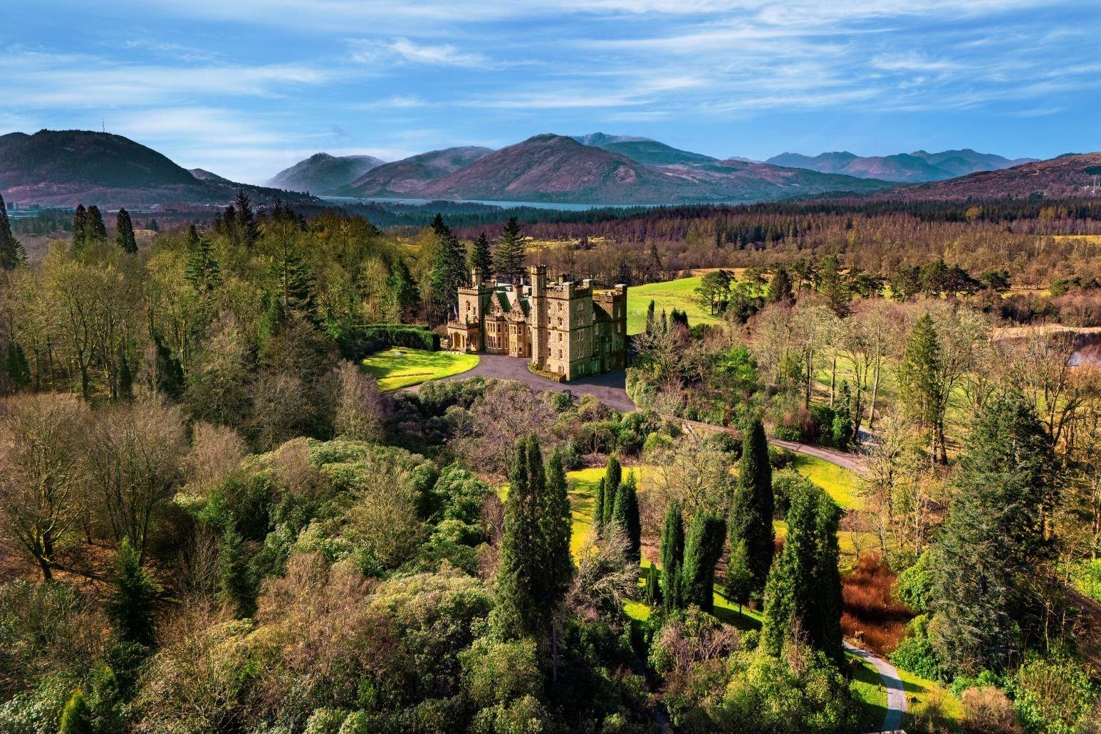 An aerial view of a turreted grey castle surrounded by lawns and trees, with a loch and layered mountains behind it under a blue sky.