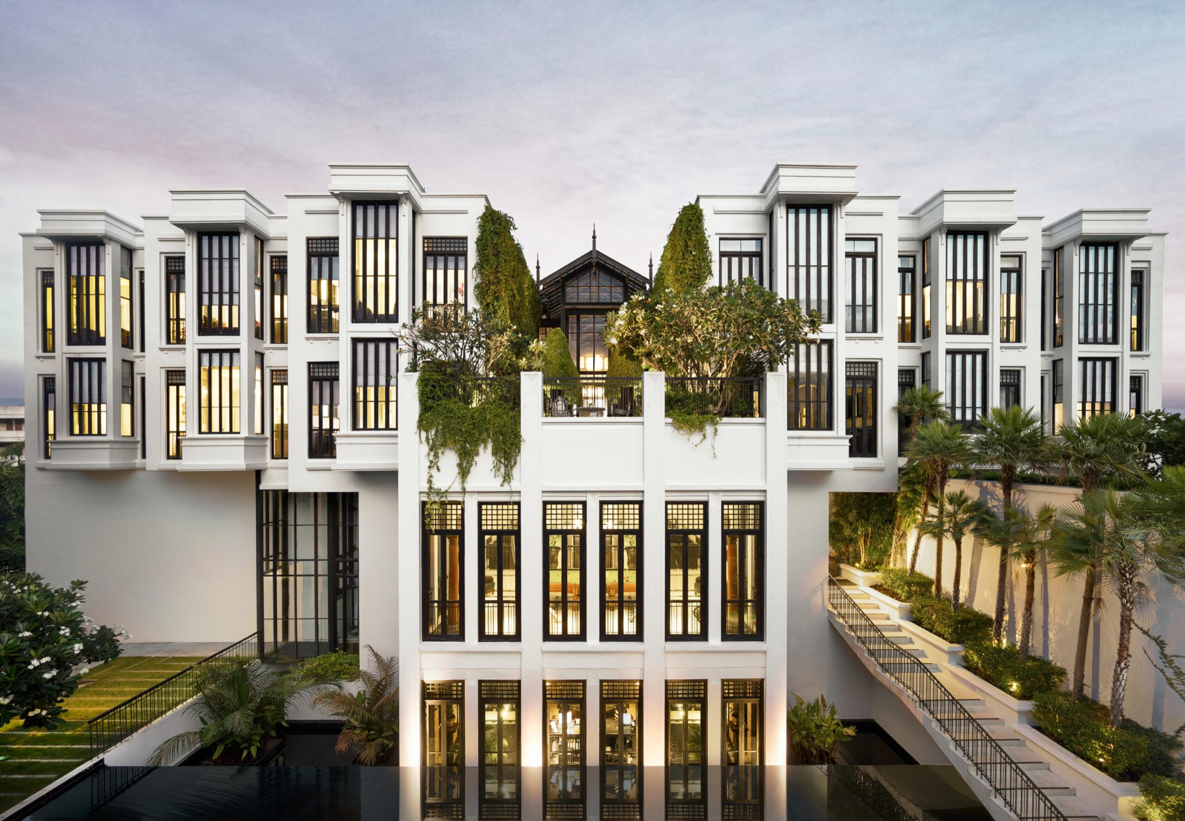 The hotel's white colonial-style building lit up at dusk, its upper floor wrapped in trees and plants, with a flight of outdoor stairs down one side and a dark reflecting pool across the front.