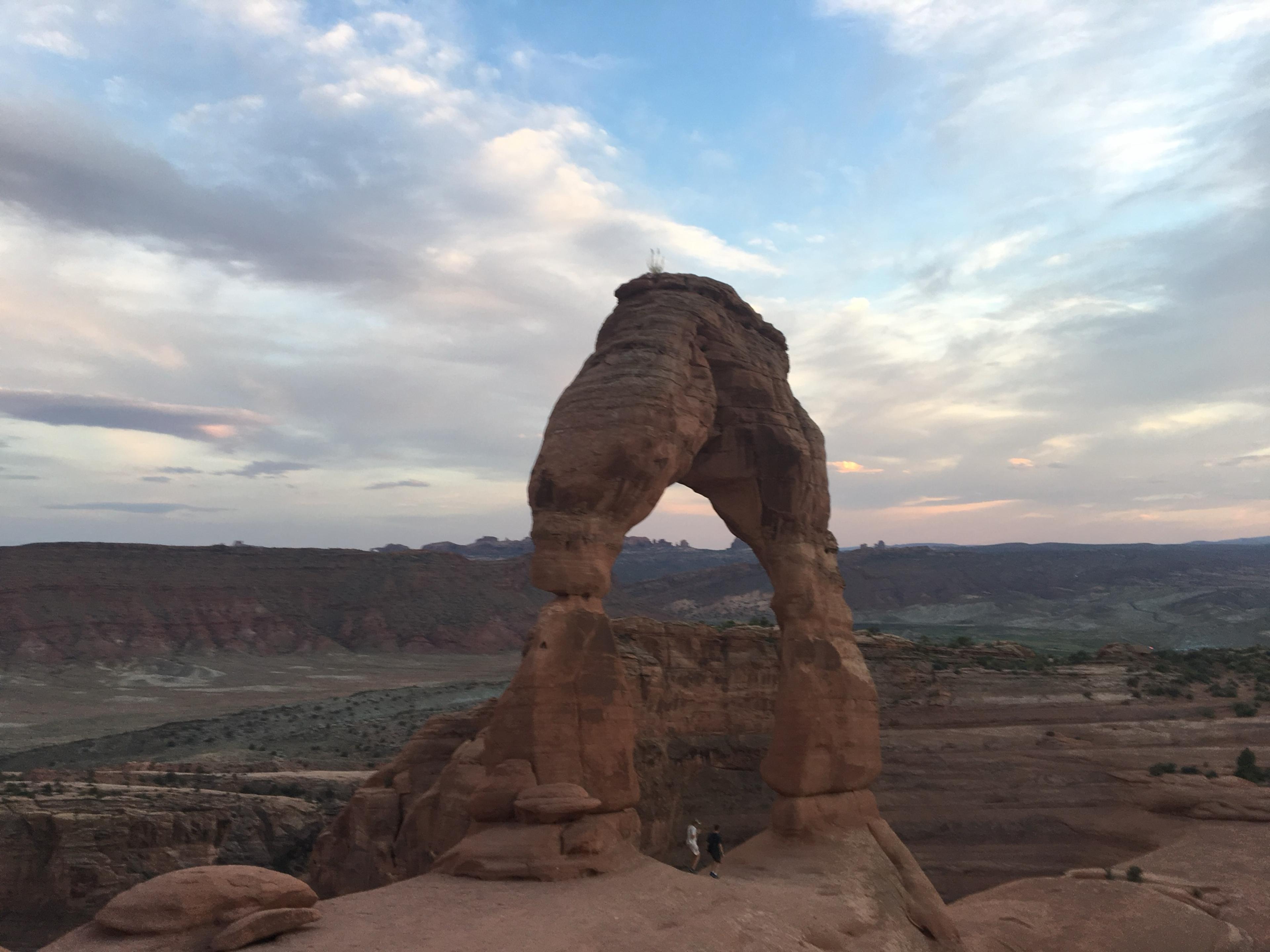 Delicate Arch, a free-standing arch of red sandstone on bare rock, in the early evening, with two people walking beneath it and the desert running away to low ridges under a pale, broken sky.