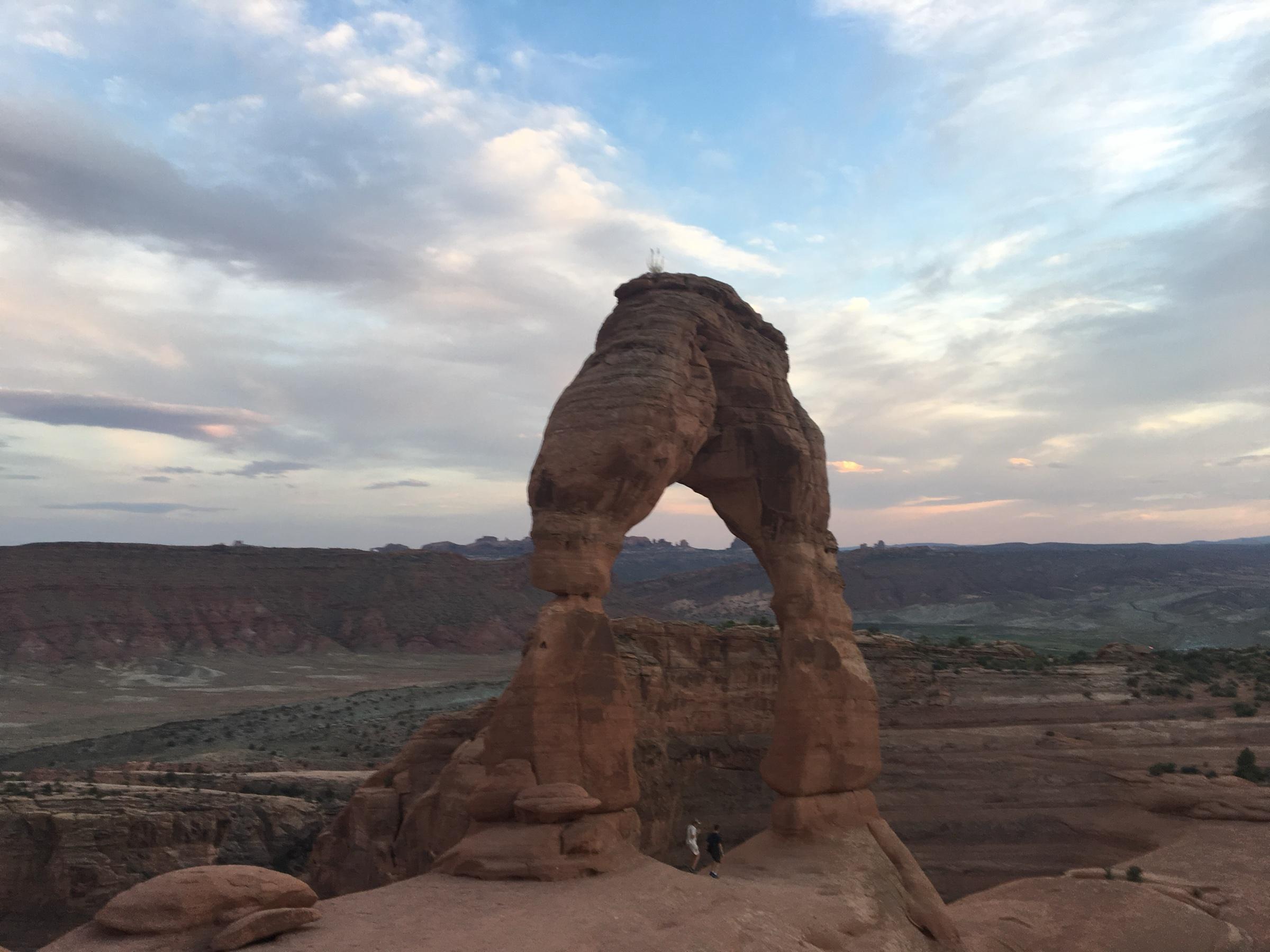 Delicate Arch, a free-standing arch of red sandstone on bare rock, in the early evening, with two people walking beneath it and the desert running away to low ridges under a pale, broken sky.