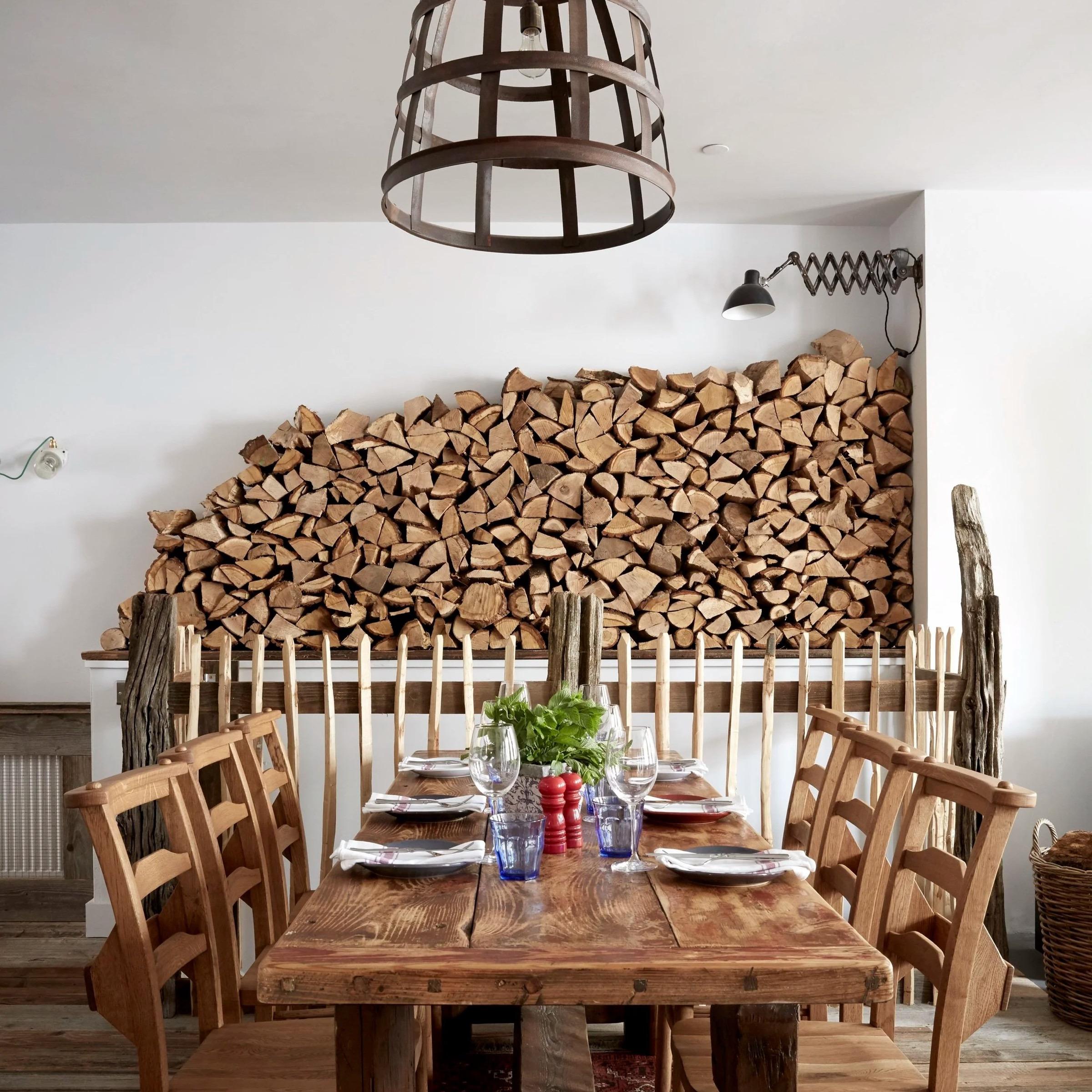 A dining table set for six beneath a large stacked log display and a woven pendant light, flanked by wooden chairs.