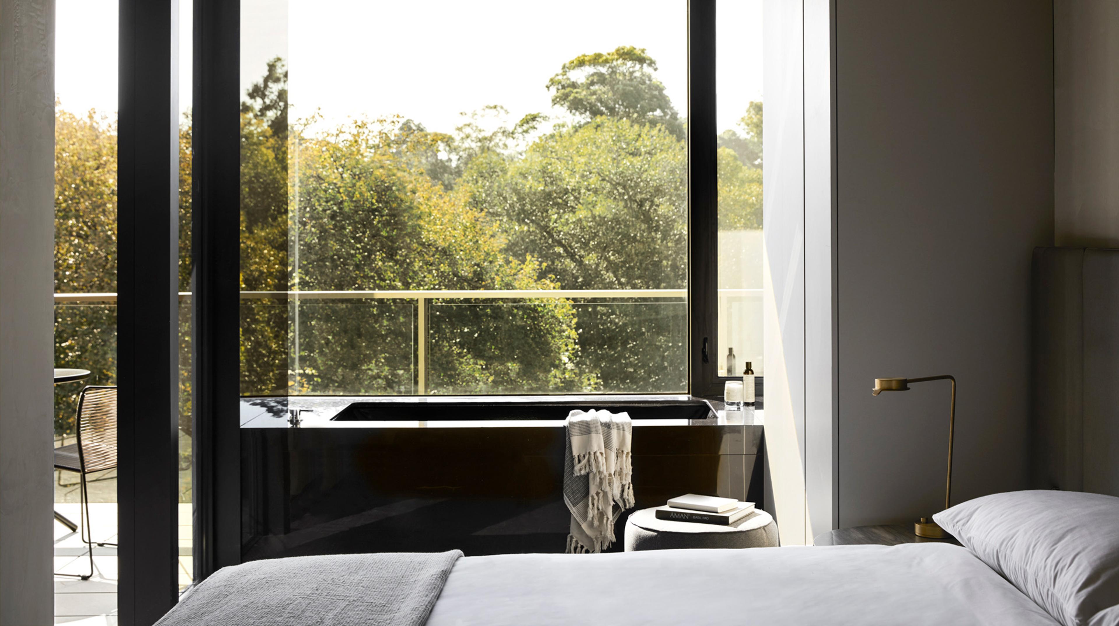 a bedroom with a black built-in bathtub beside a floor-to-ceiling window, overlooking a canopy of trees from a balcony