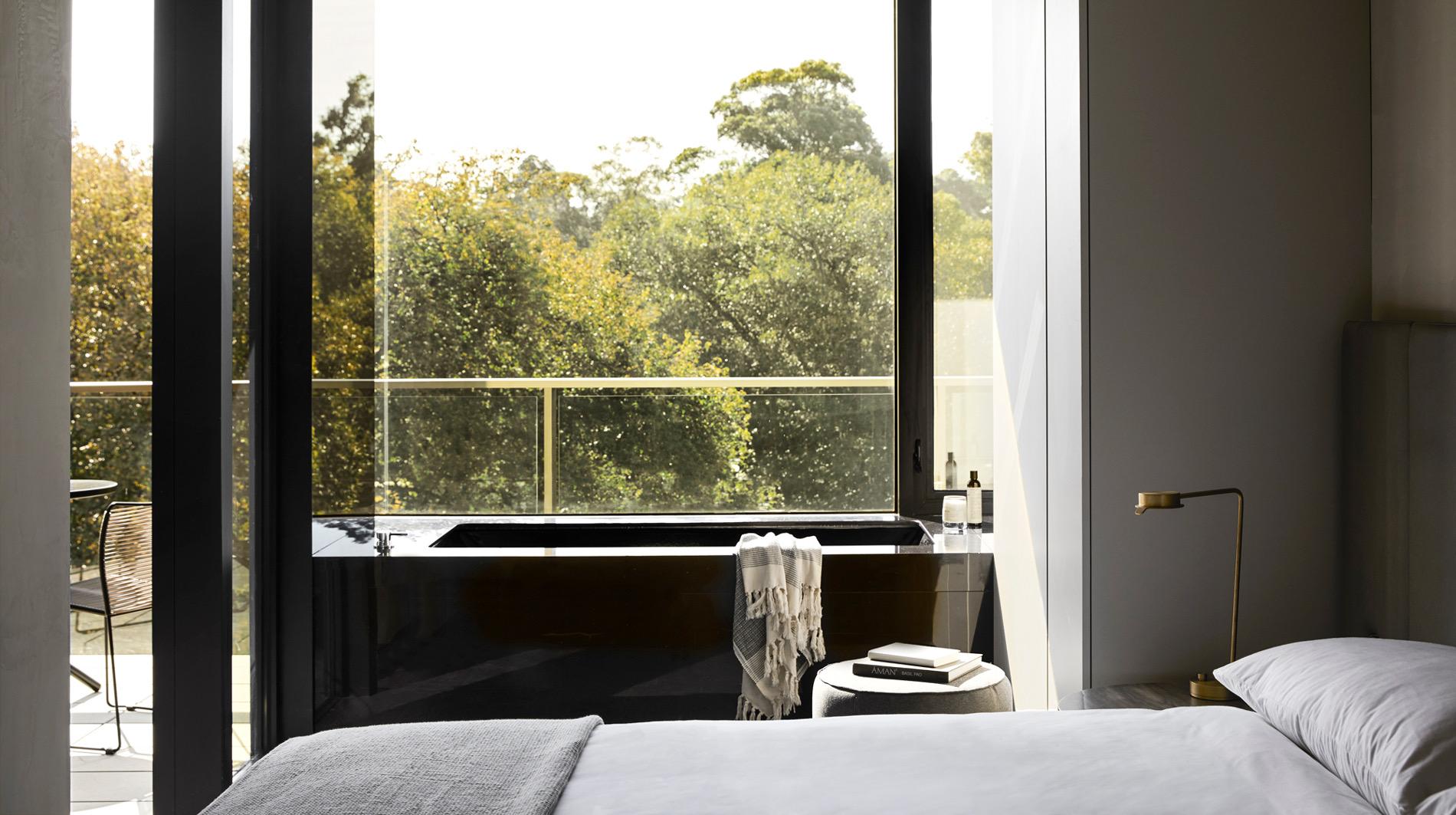 a bedroom with a black built-in bathtub beside a floor-to-ceiling window, overlooking a canopy of trees from a balcony