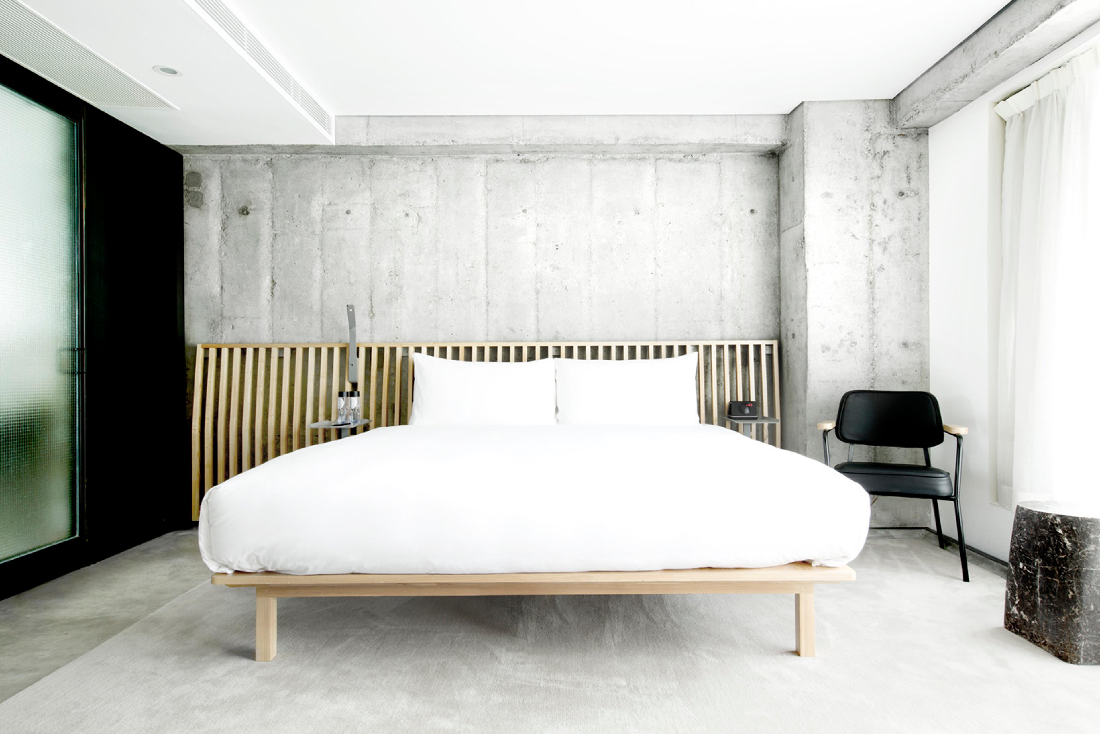 A white bed with a timber slatted headboard against a bare concrete wall, with a black chair and sheer curtain beside it.