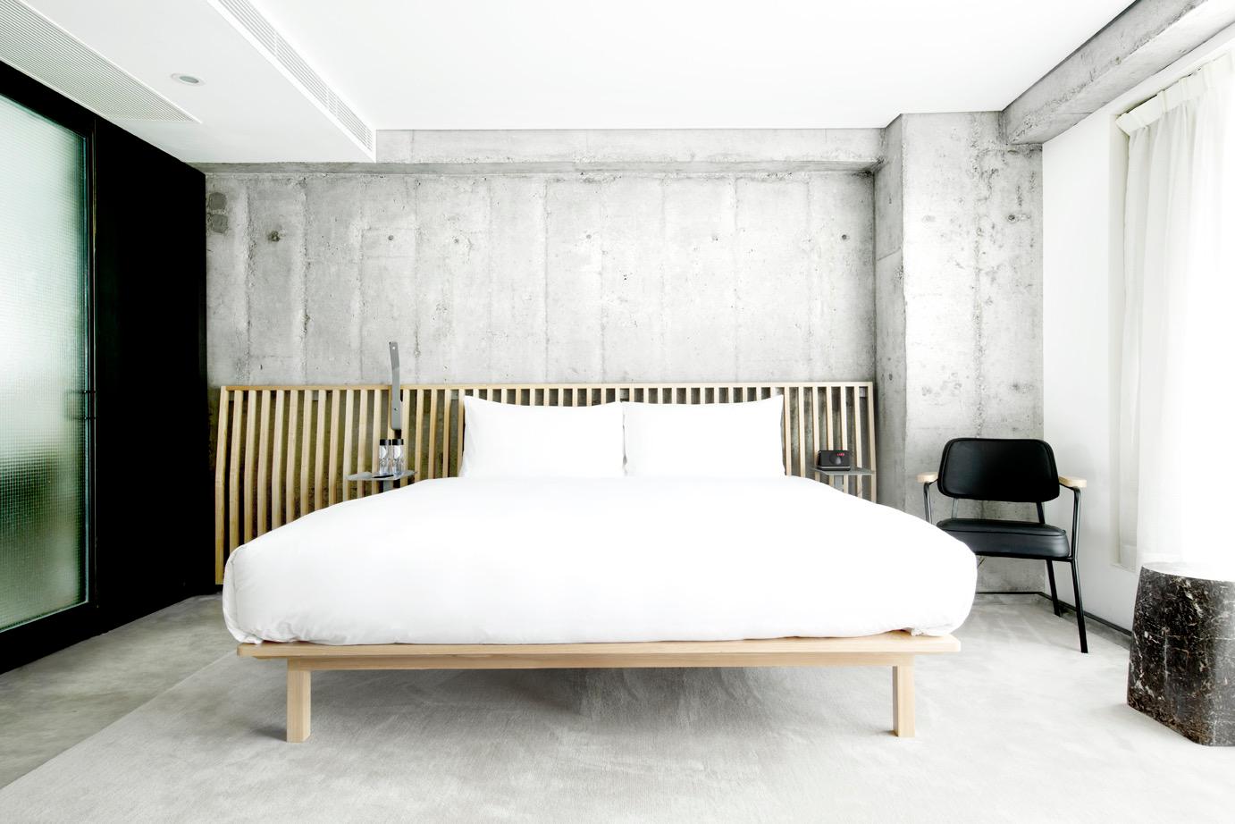 A white bed with a timber slatted headboard against a bare concrete wall, with a black chair and sheer curtain beside it.