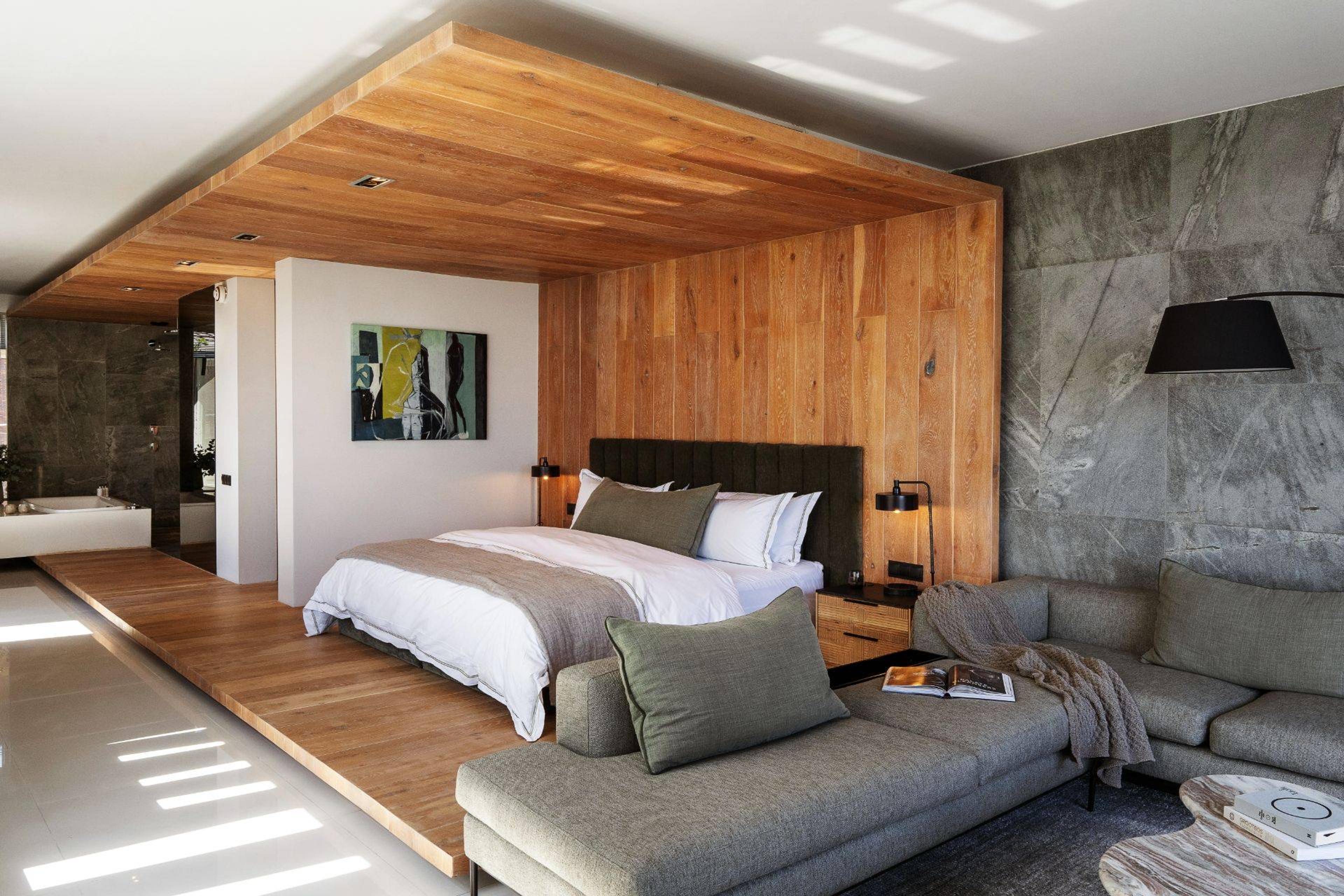 A suite with a bed, a grey sofa, a freestanding bathtub visible beyond a partition, and an abstract painting on the wall.