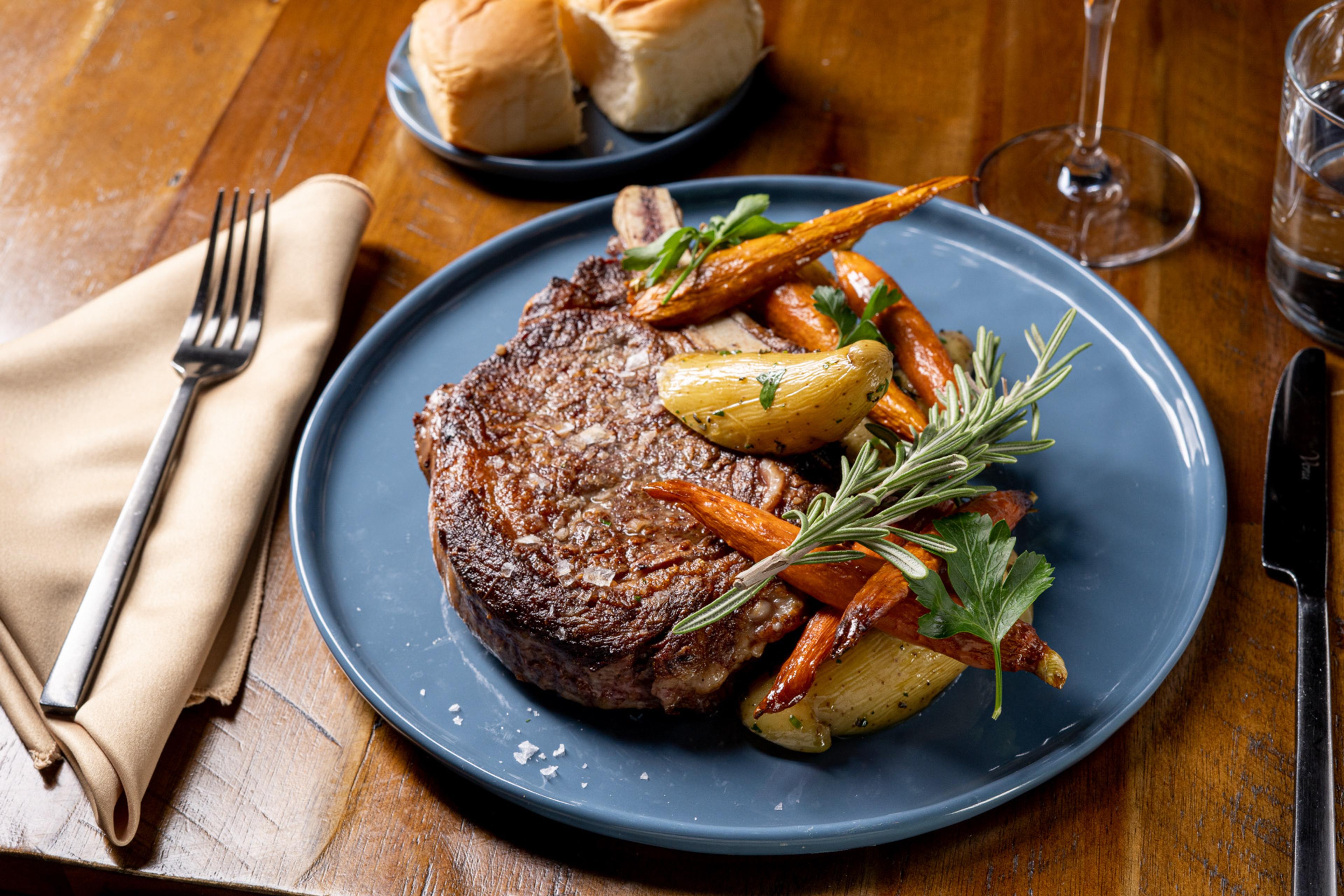 A steak with roasted carrots, baby potatoes and rosemary sits on a blue plate, with bread rolls, a fork and a knife on a wooden table.