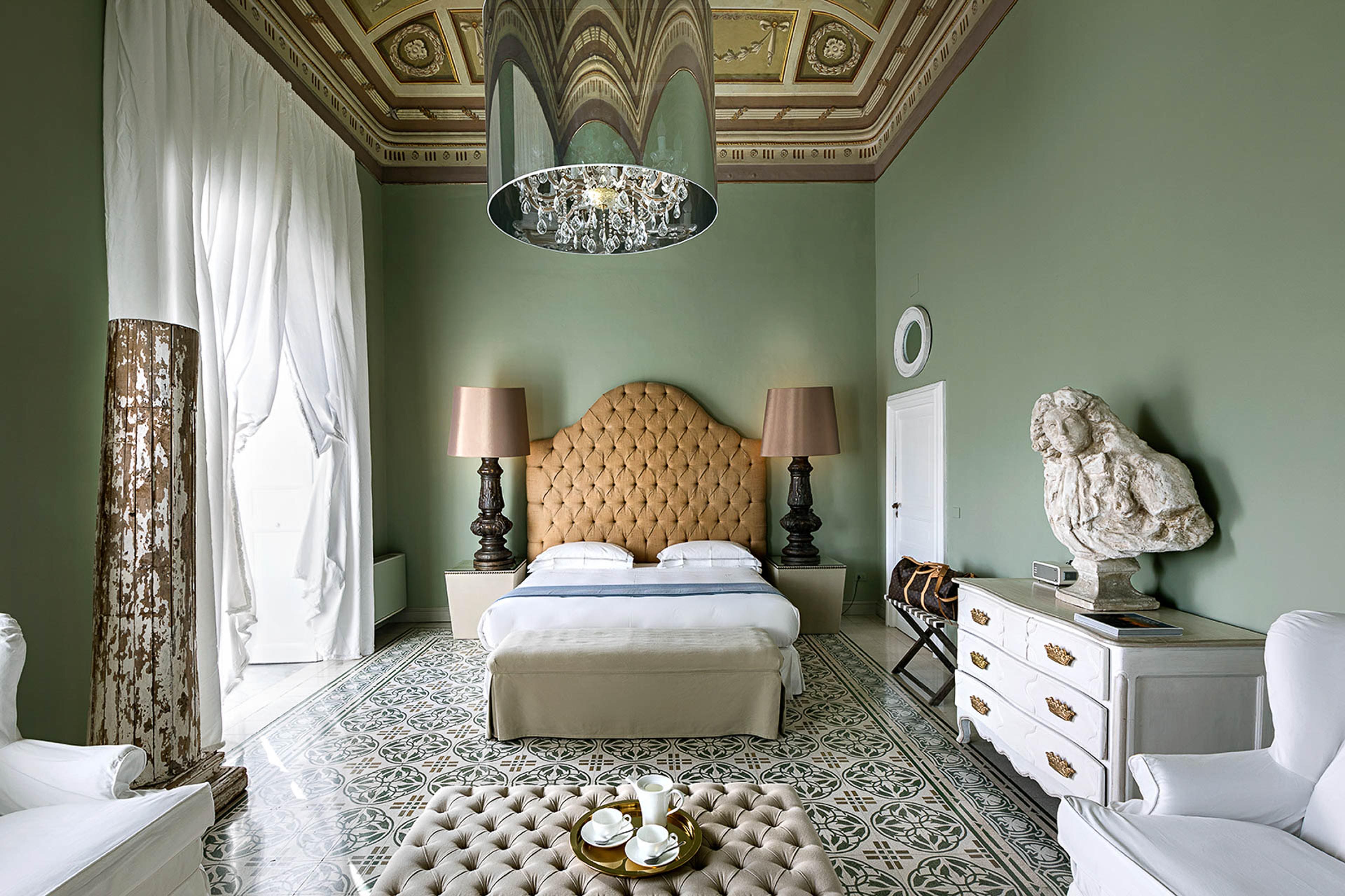 A green-walled bedroom with an ornately painted ceiling, a tufted bed, a stone sculpted bust on a chest of drawers, and a patterned tiled floor.