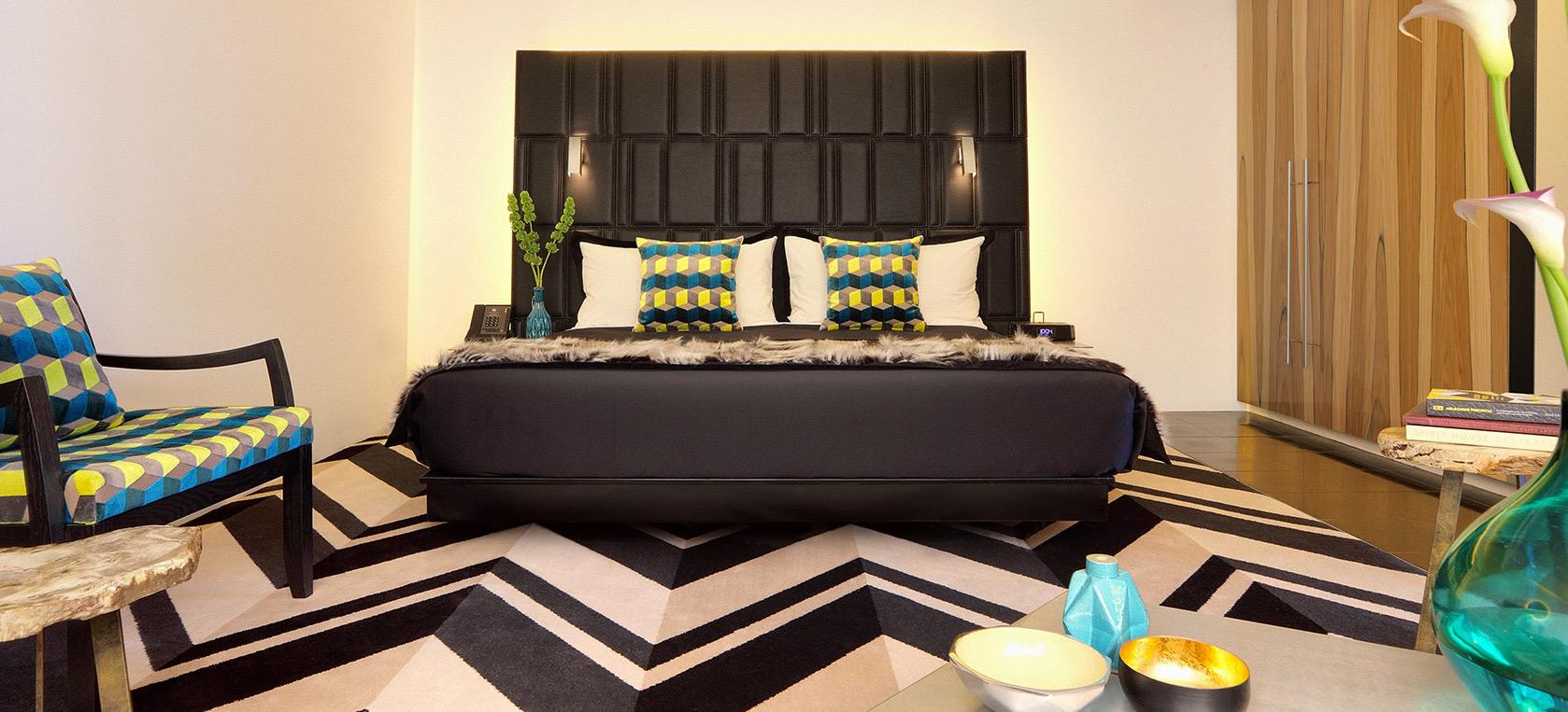 A bedroom with a black tufted leather headboard, teal and yellow patterned cushions, and a matching armchair on a chevron rug.