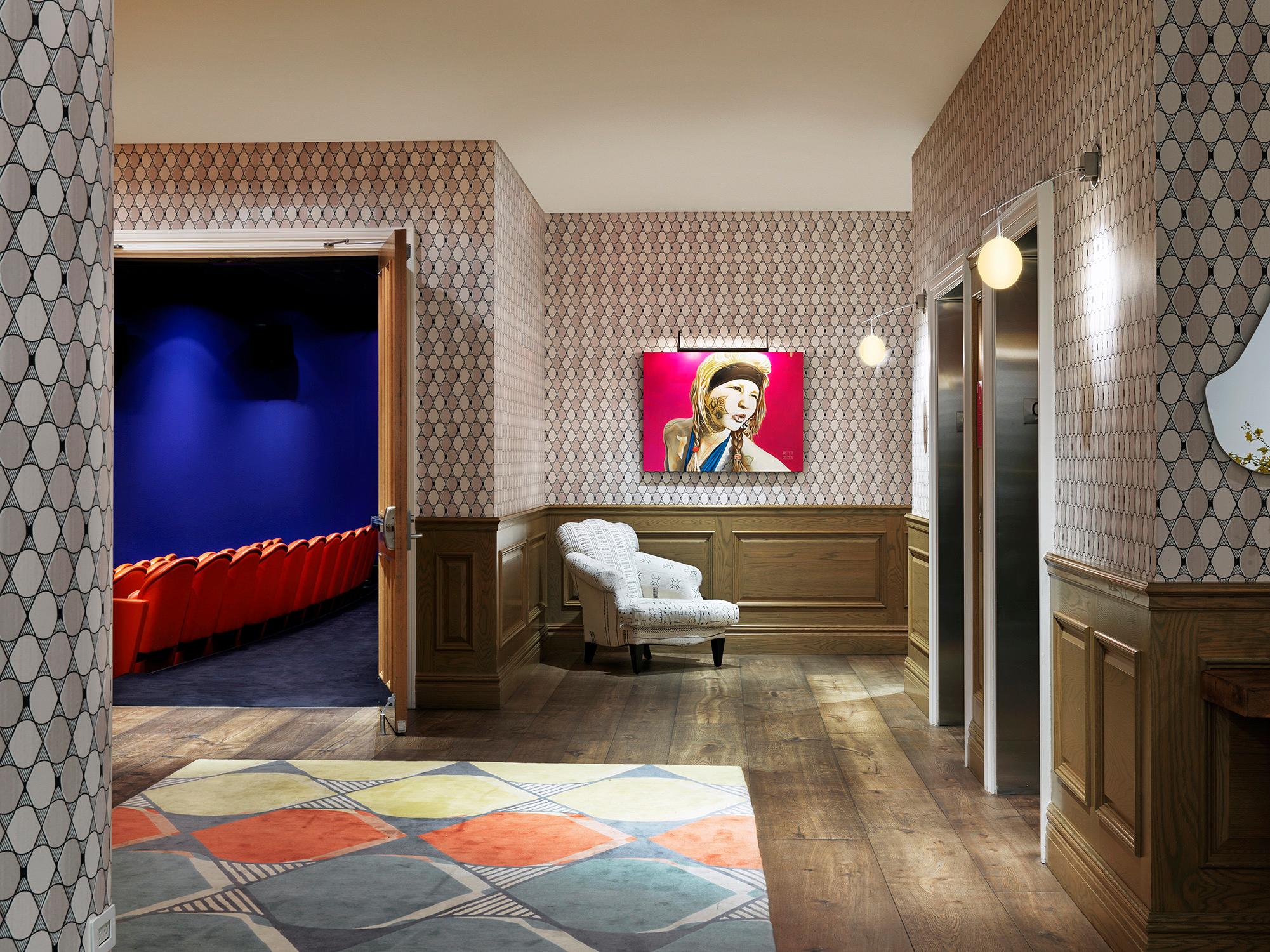 A corridor with patterned wallpaper, a portrait of a woman on the wall, an armchair, and an open door showing rows of red seats in a small screening room.