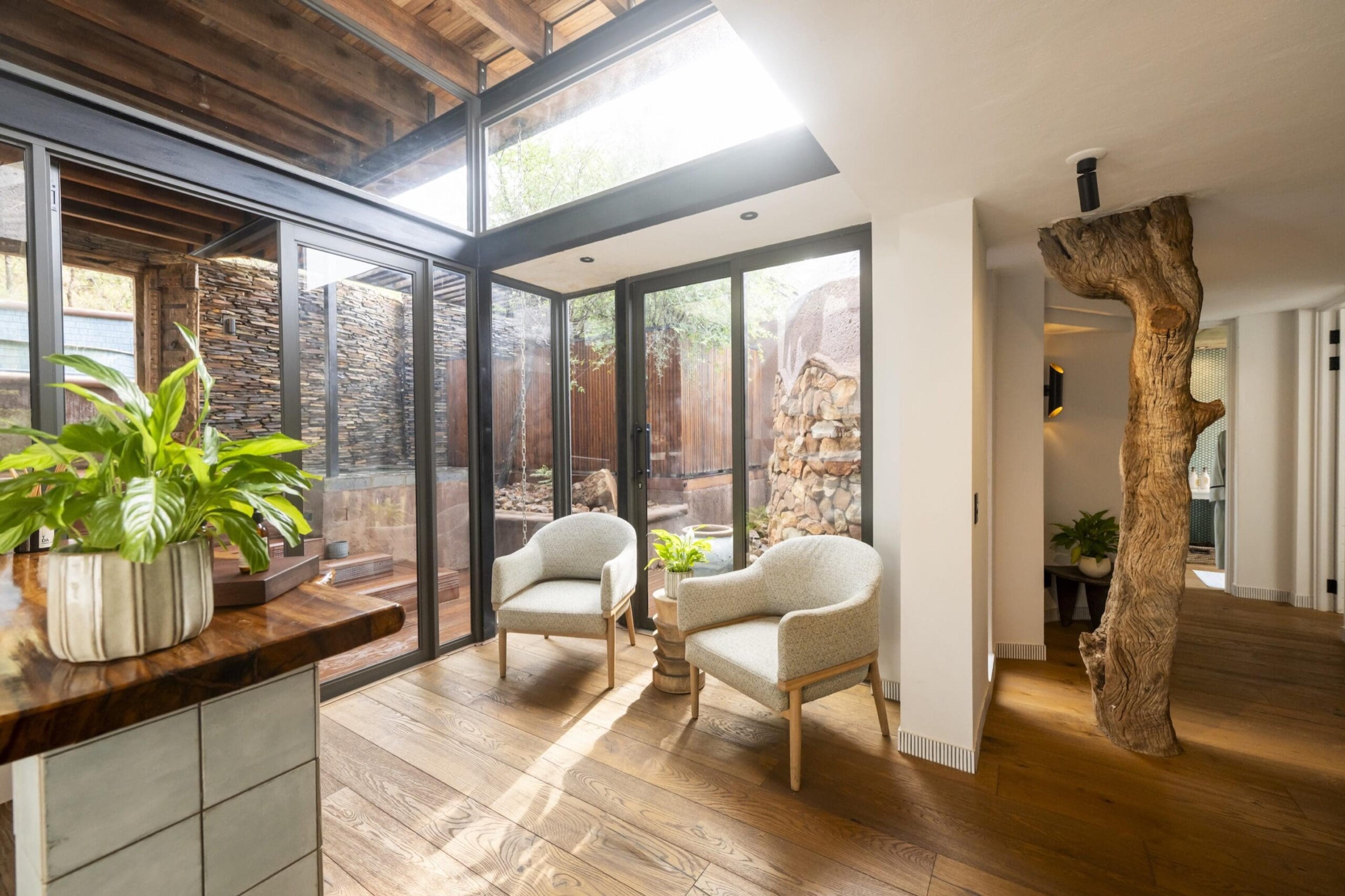A sitting area with two armchairs and a low table, a potted plant on a wooden counter, stone and timber walls, and a bare tree trunk used as a structural feature