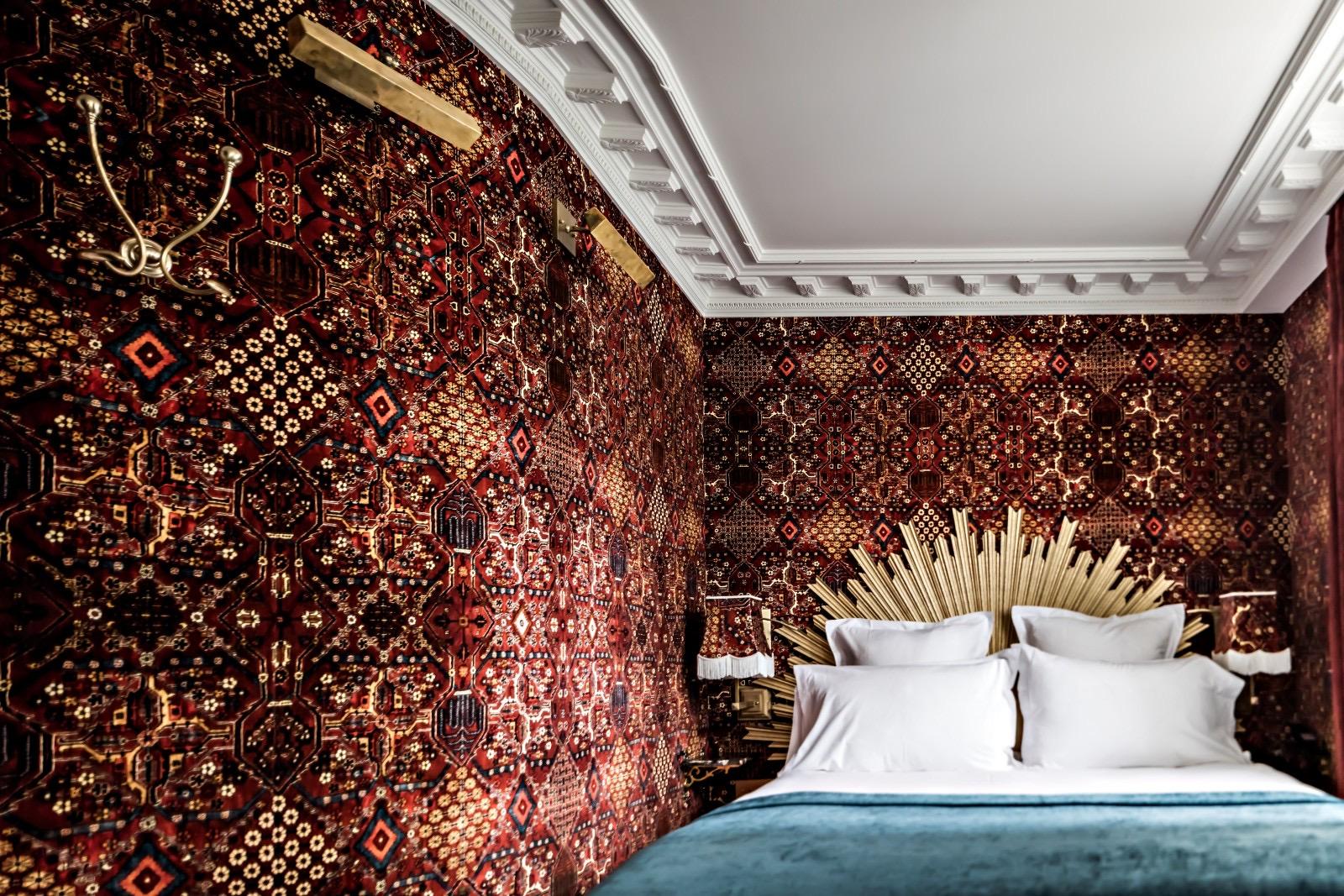 A bed with white pillows and a teal velvet throw sits below a gold sunburst headboard, against dark red patterned wallpaper that wraps around a curved corner.