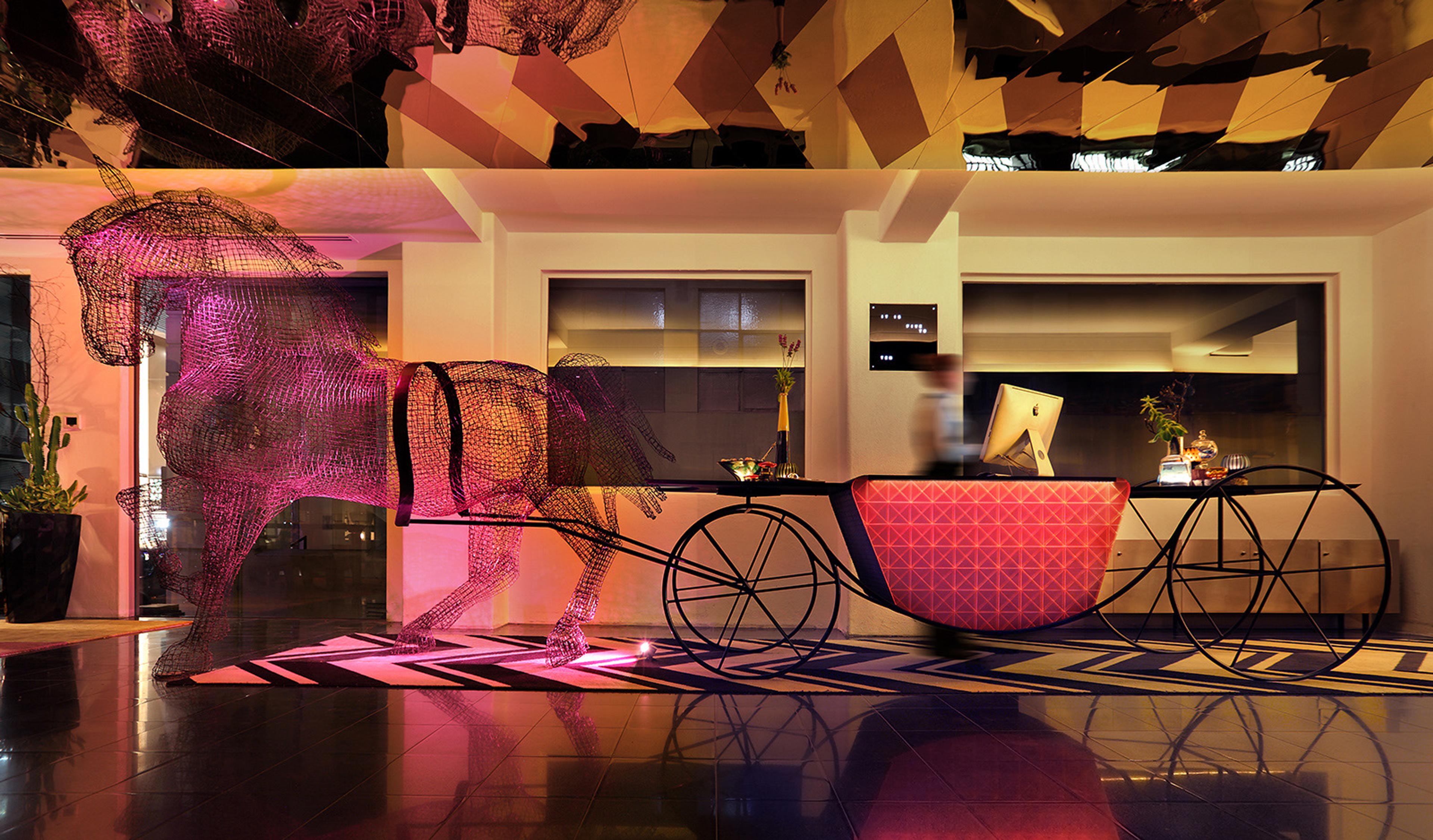 A hotel lobby at night with a wire sculpture of a horse and carriage used as a reception desk, lit in pink and gold, with a staff member blurred in motion behind it.